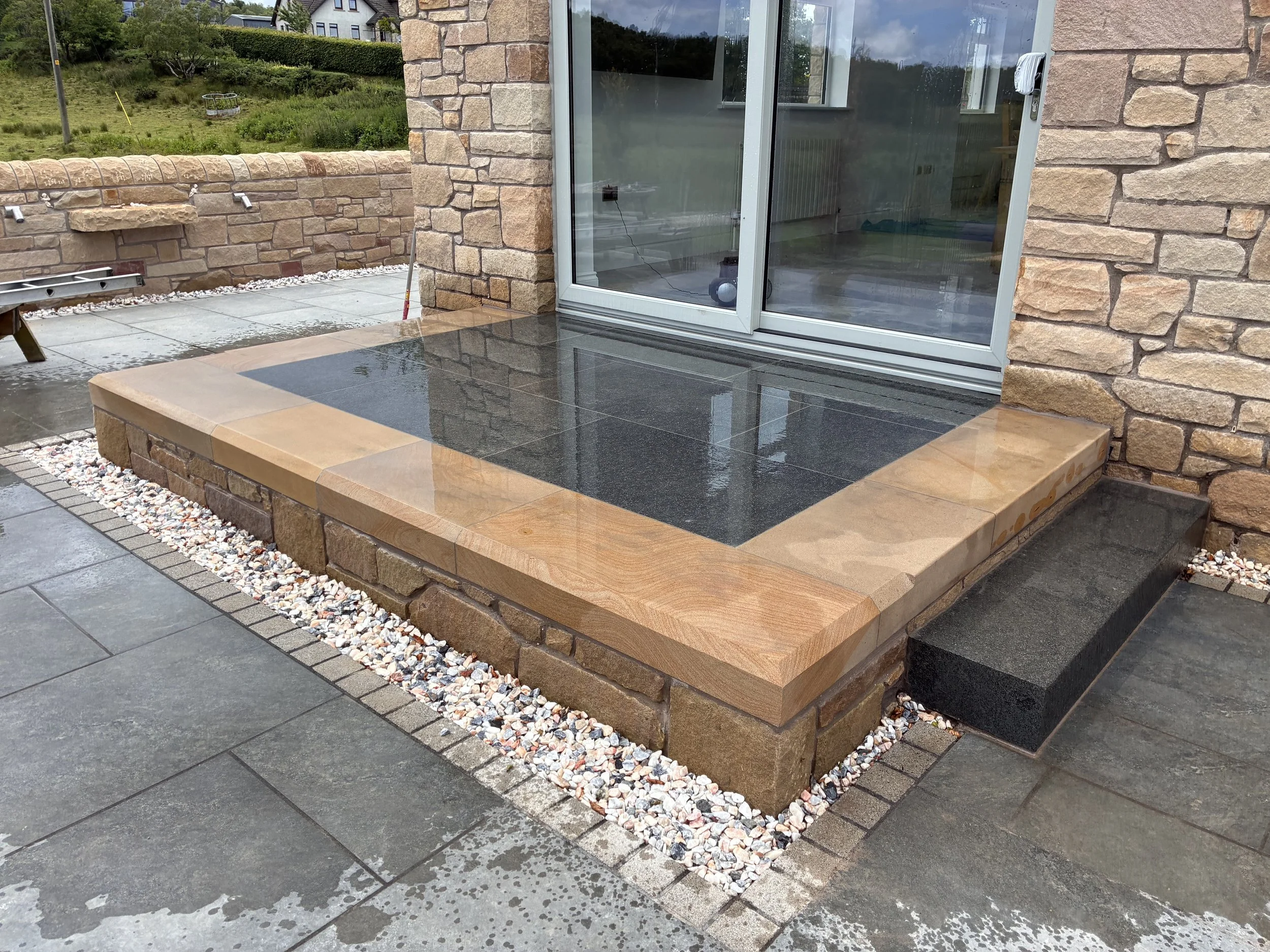 The image shows part of a patio area with a black tiled surface, attached to a building with a stone brick exterior. There is a small step of beige stone leading down to a darker tiled outdoor floor. To the left, a stone wall with a small shelf, alon