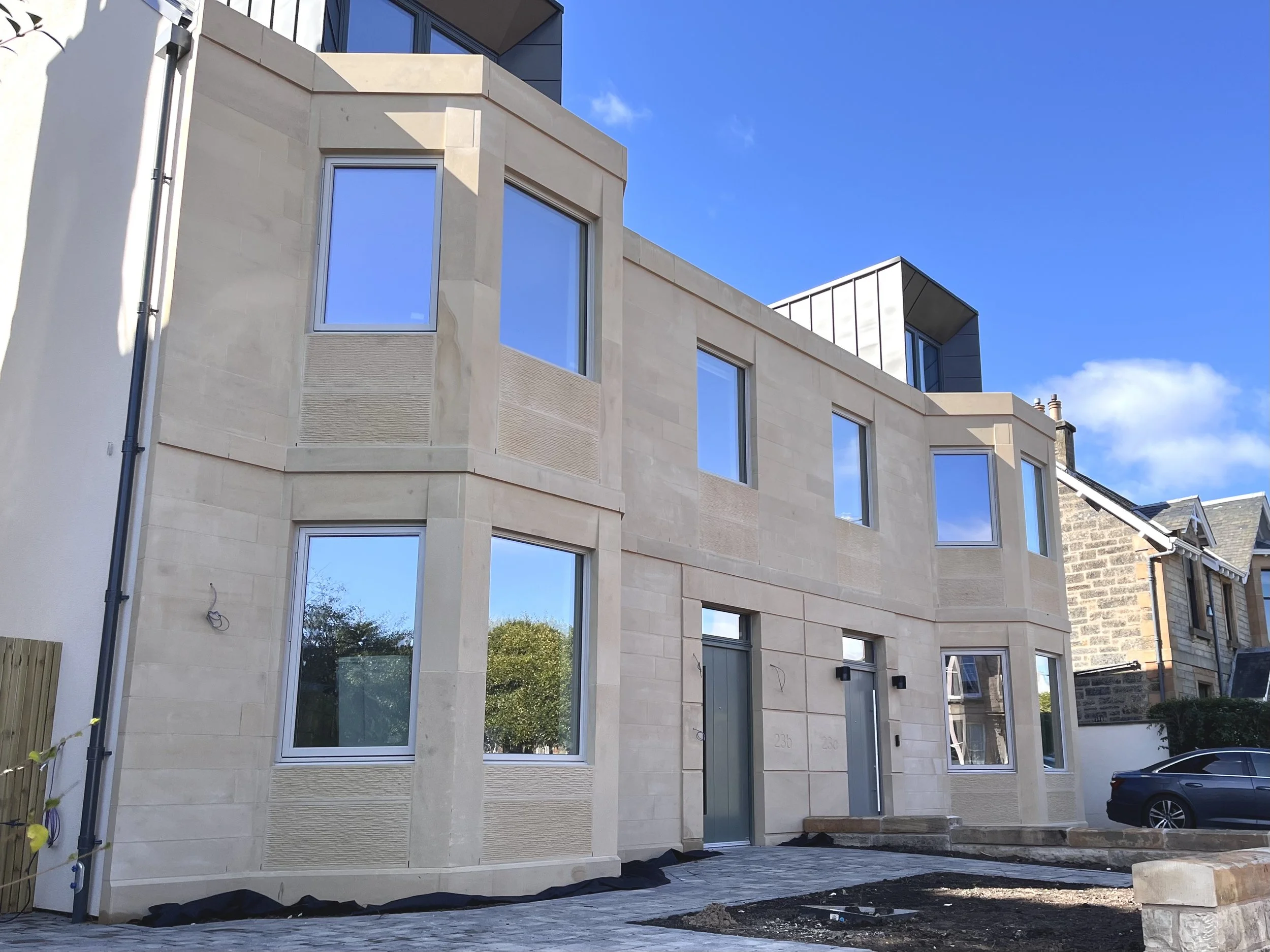 Modern multi-story residential building with large windows, beige stone exterior, and a small courtyard area with paving stones and recently planted garden beds, under a clear blue sky.