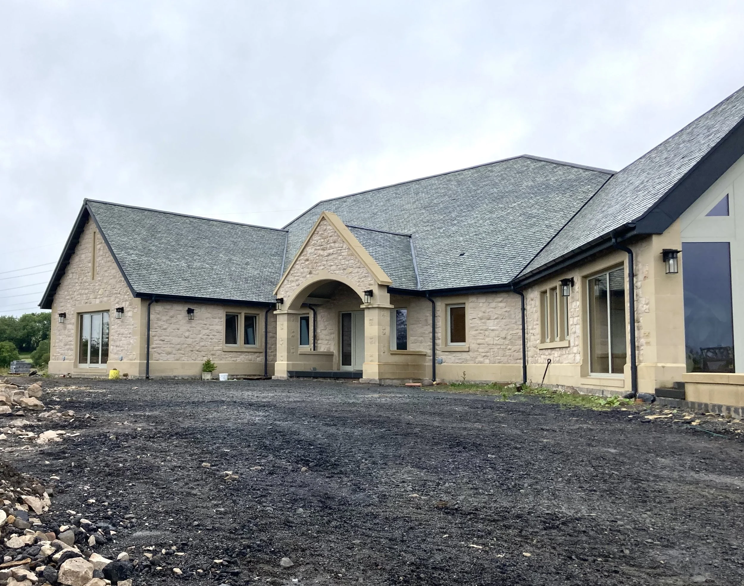 Newly constructed house with stone and siding exterior, multiple gabled roofs, large windows, and a gravel driveway, under an overcast sky.