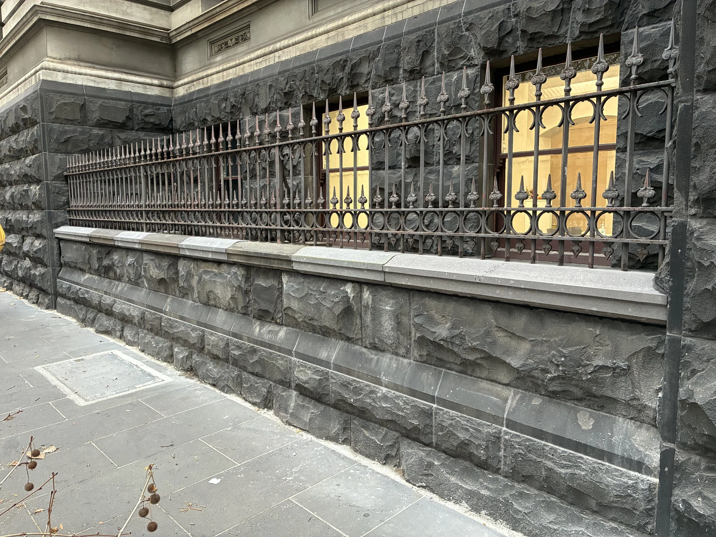 A stone building with a metal fence and a window with a yellow curtain, sidewalk with some dried plants near the curb.