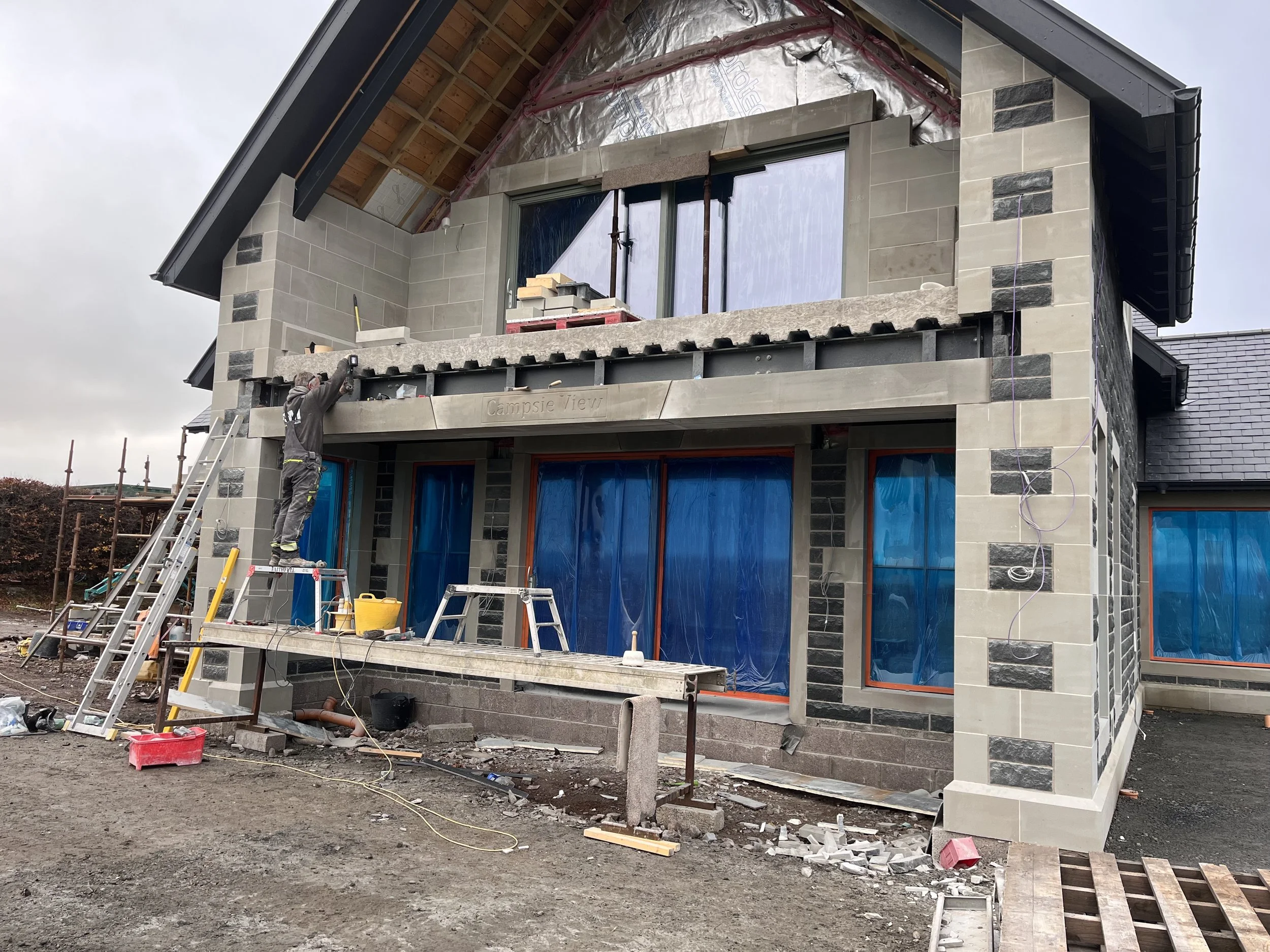 Construction site of a modern house with partially finished stone exterior, workers, scaffolding, tools, and materials visible.