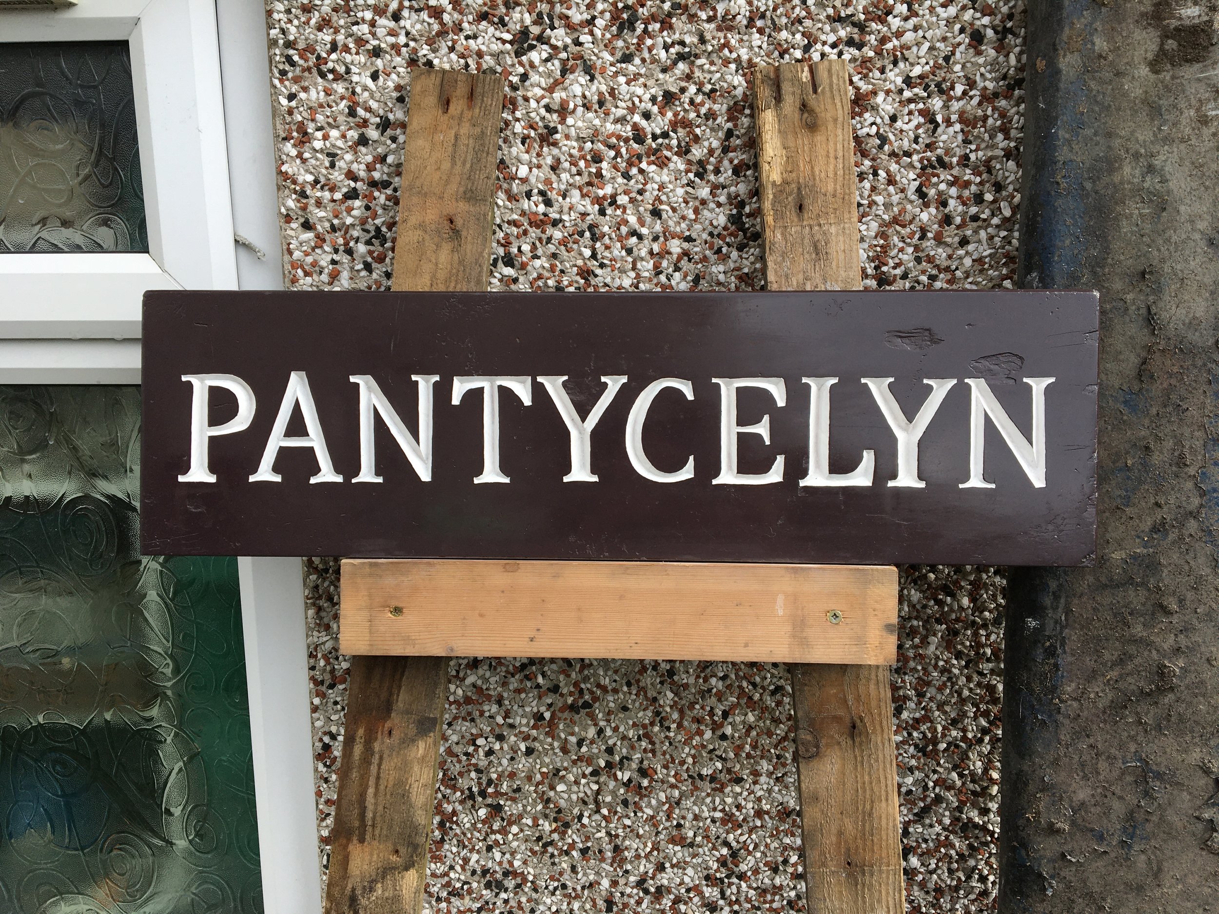 Wooden planks and a sign with the word 'PANTYCELYN' on a rocky ground outside a building with a glass window and white framing.