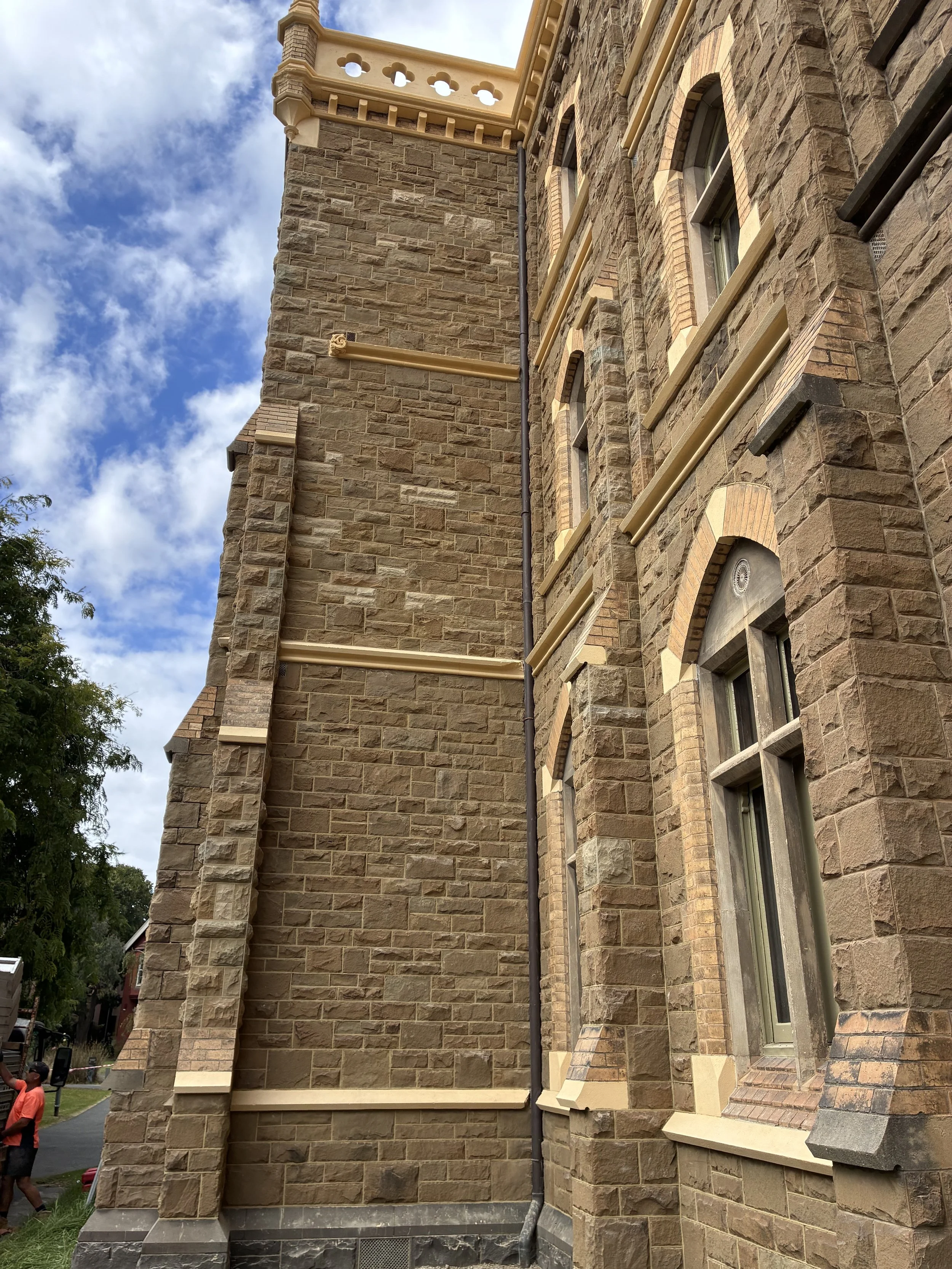 The corner of a historic stone building with arched windows and decorative trim, under a partly cloudy sky. A black downspout runs along the building's corner.