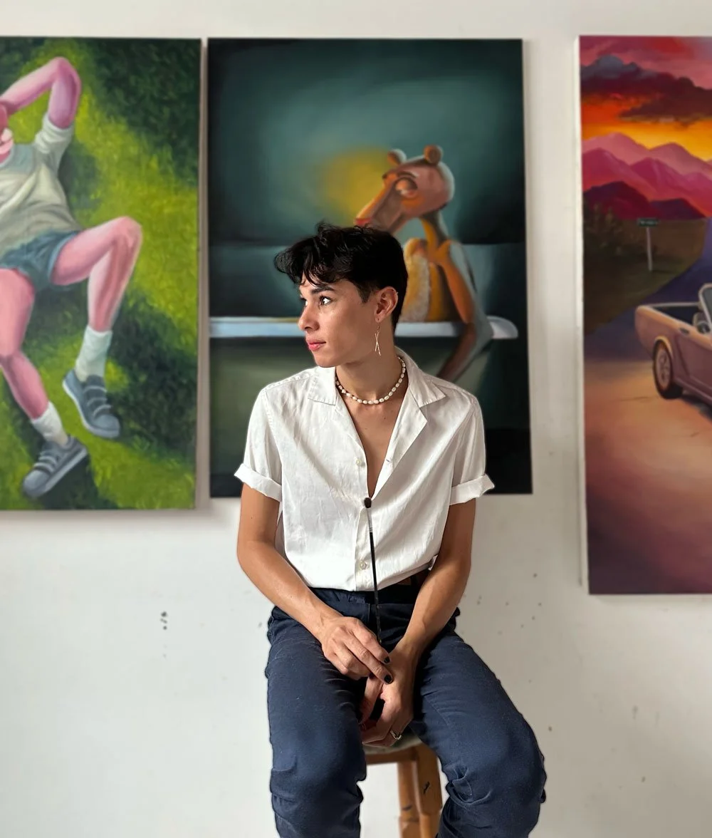 Mujer con prendas blancas y pantalones oscuros en una galería de arte con pinturas coloridas en la pared.