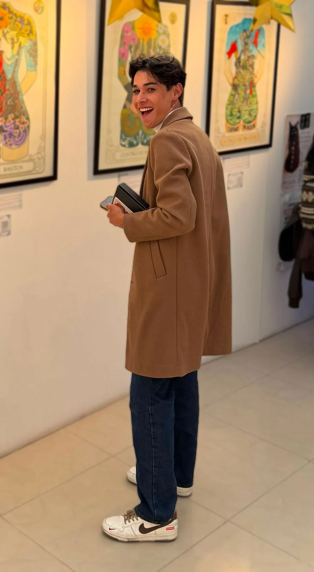 Joven sonriendo en una galería de arte, con cuadros coloridos en la pared y decoraciones doradas en el techo.