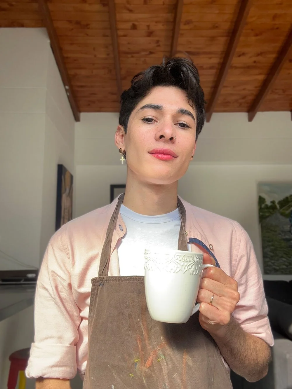 Joven con delgado rostro, cabello corto y oscuro, sosteniendo una taza blanca con detalles en relieve, vistiendo camiseta blanca y delantal marrón, en una cocina con techo de madera.