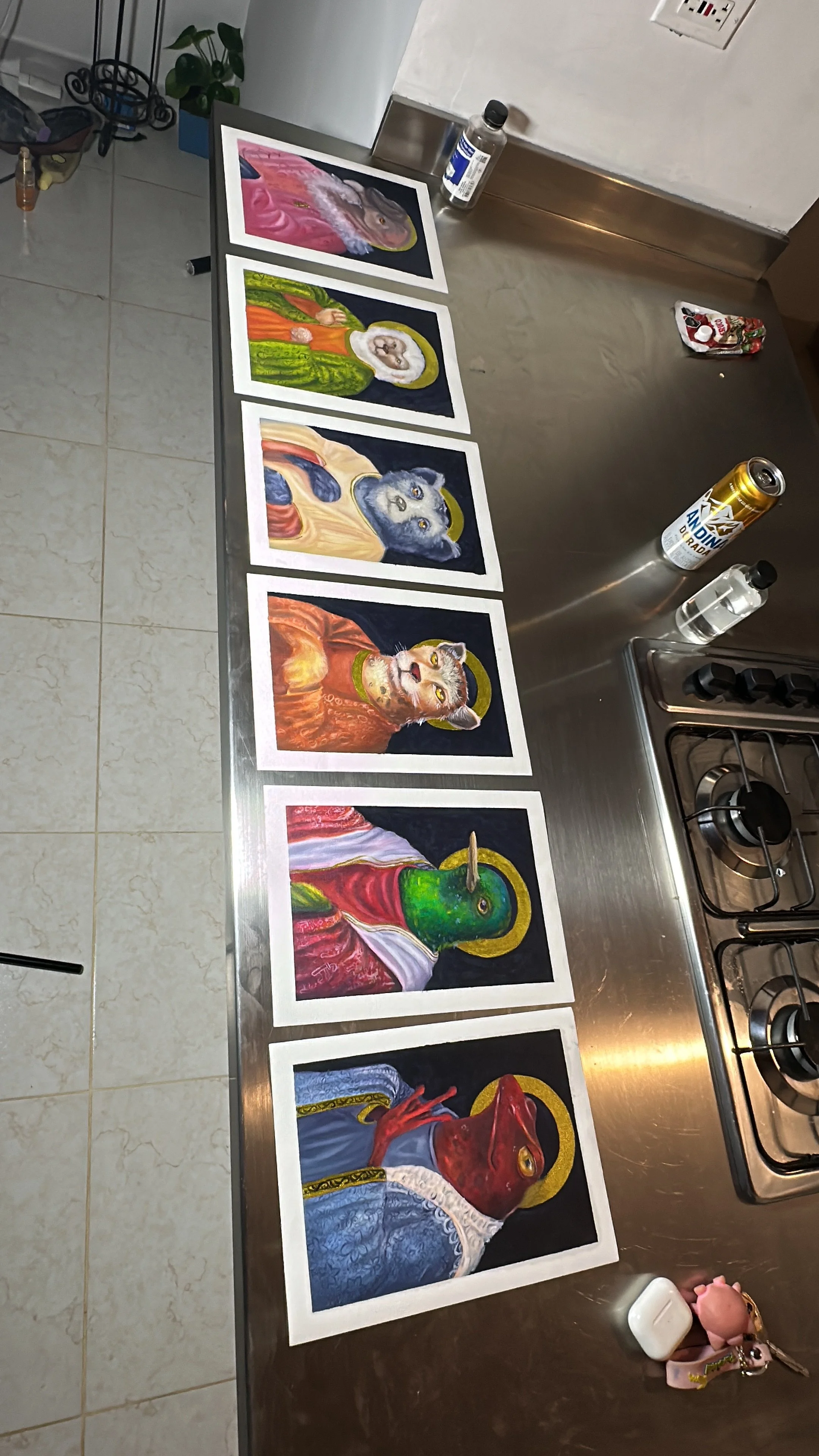 Seis pinturas de animales con halos dorados en fondo oscuro, colocadas sobre una mesa de cocina de acero inoxidable.