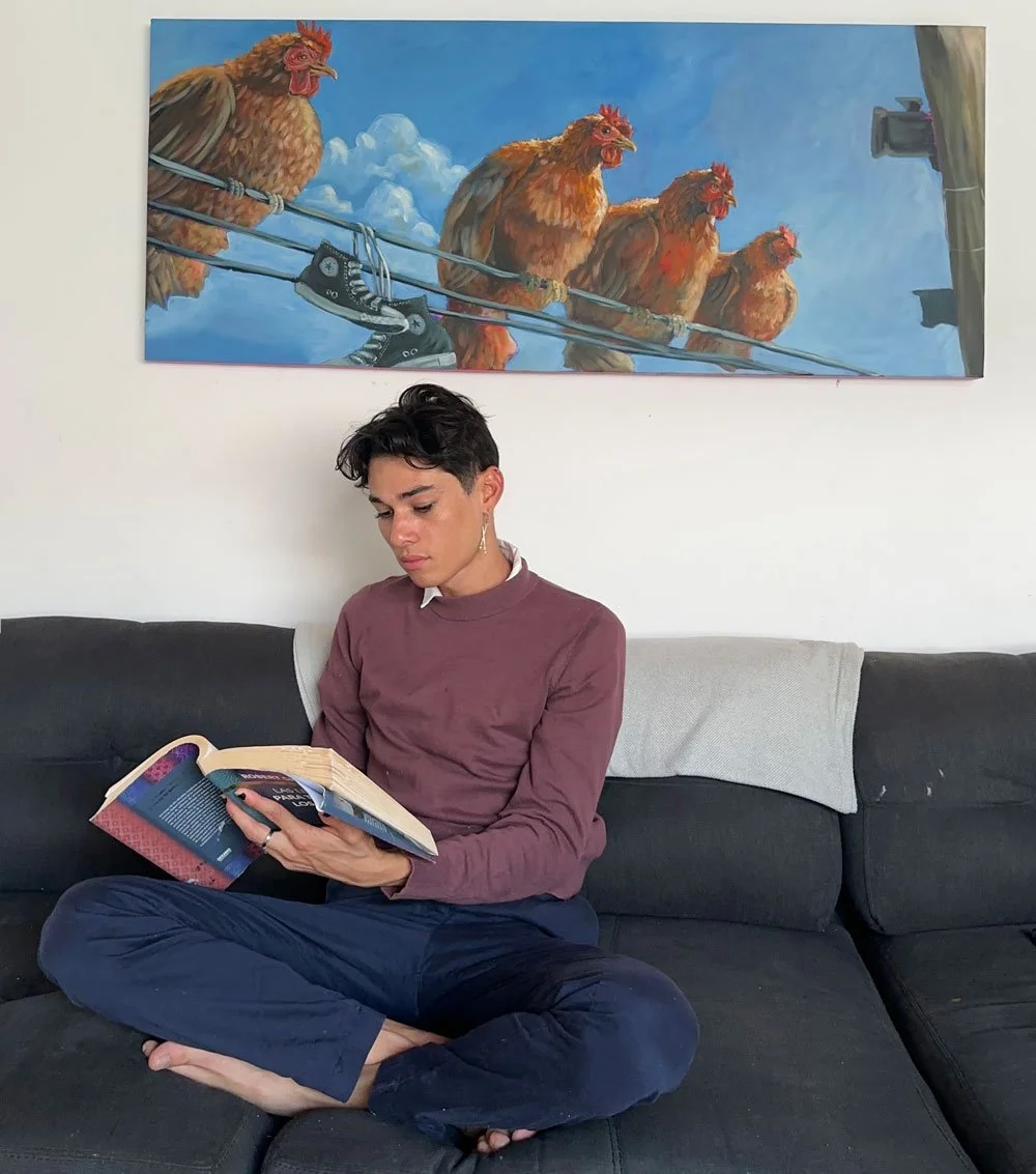 Una persona sentada en un sofá leyendo un libro en una sala con un cuadro de gallinas en el fondo.