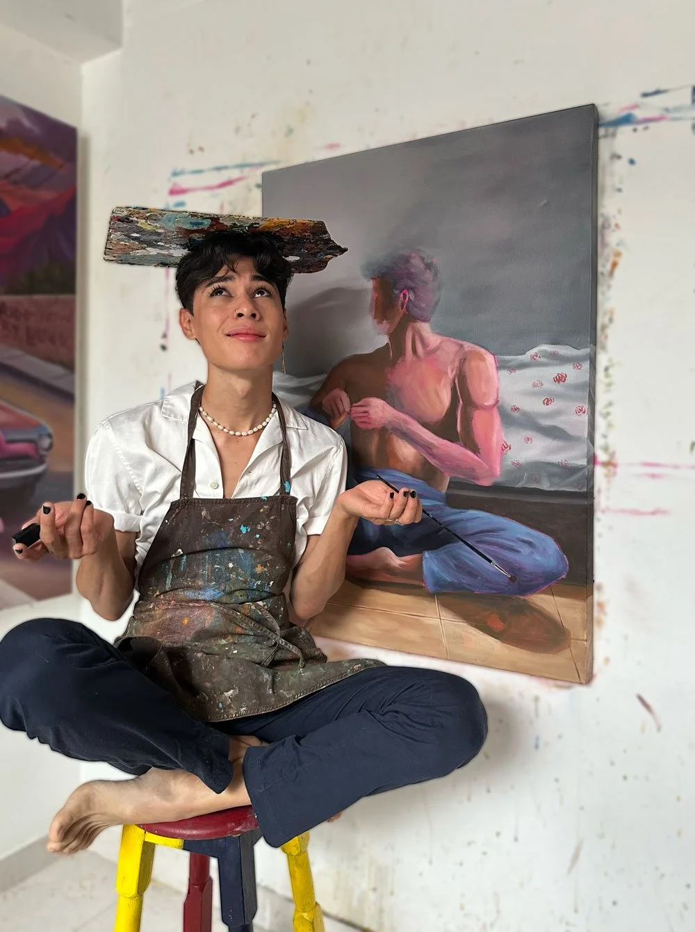 Una Joven artista con delantal manchado de pintura, sentado en un banquito con las piernas cruzadas en una sala de arte, posando junto a su pintura de un hombre desnudo en una postura similar.