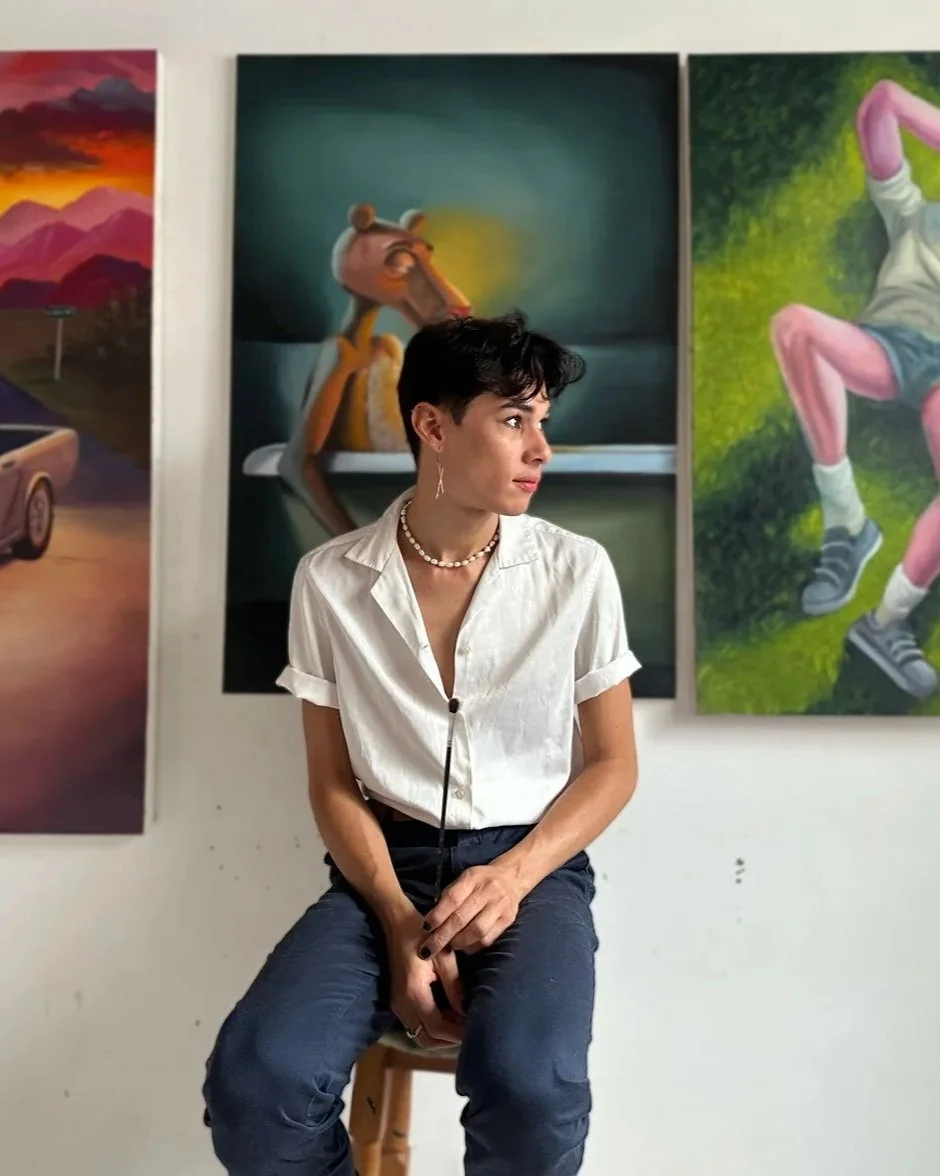 Un joven sentado en un taburete en una galería de arte, con pinturas en la pared detrás de ella que incluyen una escena de paisaje y personajes de dibujos animados.