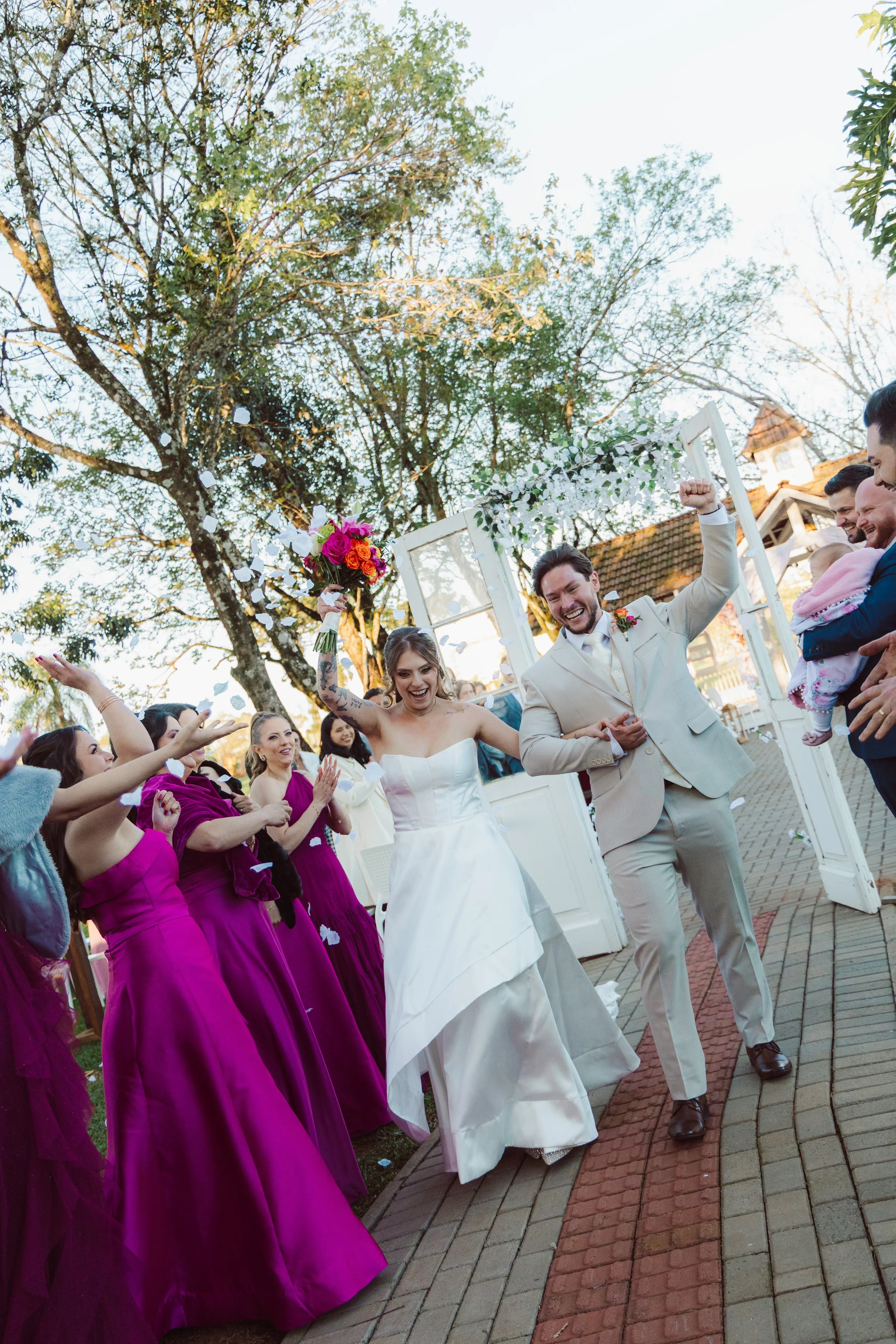 A bride and groom celebrating at their wedding, surrounded by friends and family. The bride is wearing a white wedding gown and holding a colorful bouquet, while the groom is dressed in a light-colored suit with a boutonniere. They are smiling and raising their hands in happiness, with a backdrop of trees and a white outdoor structure.