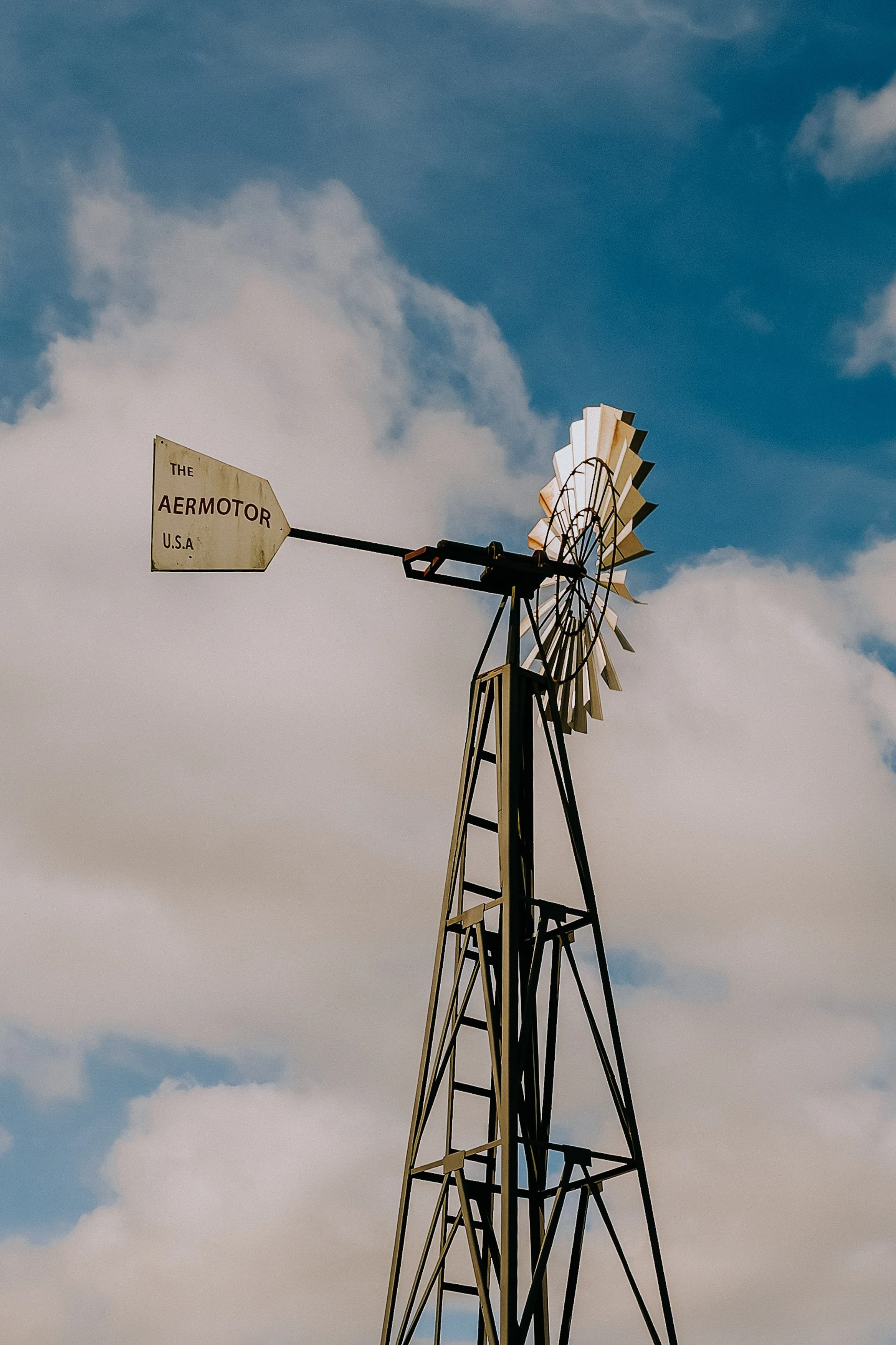 A vintage windmill with a white tail and blades against a partly cloudy blue sky.