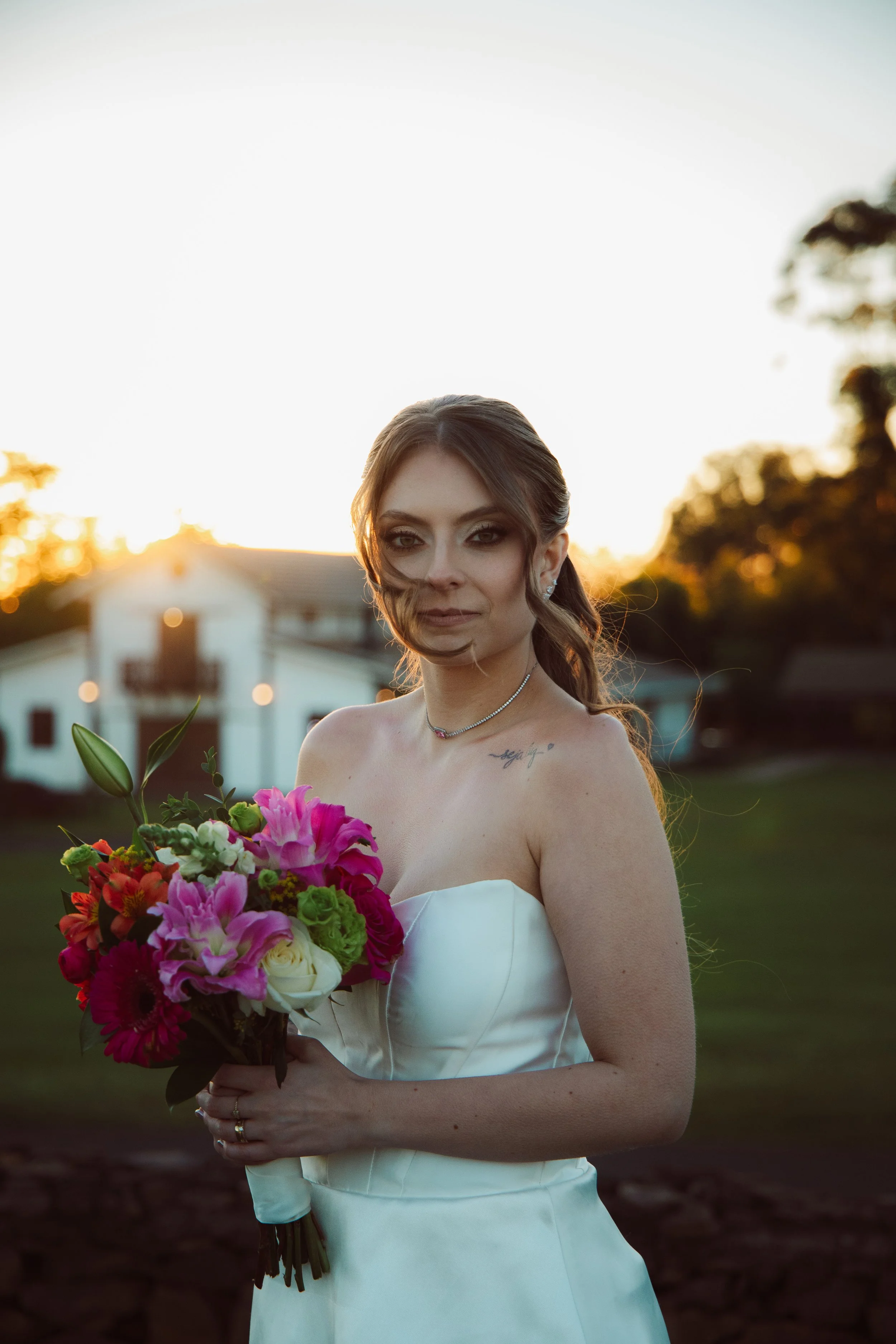 A woman in a white strapless wedding dress holding a bouquet of pink, red, and white flowers during sunset with a house and trees in the background.
