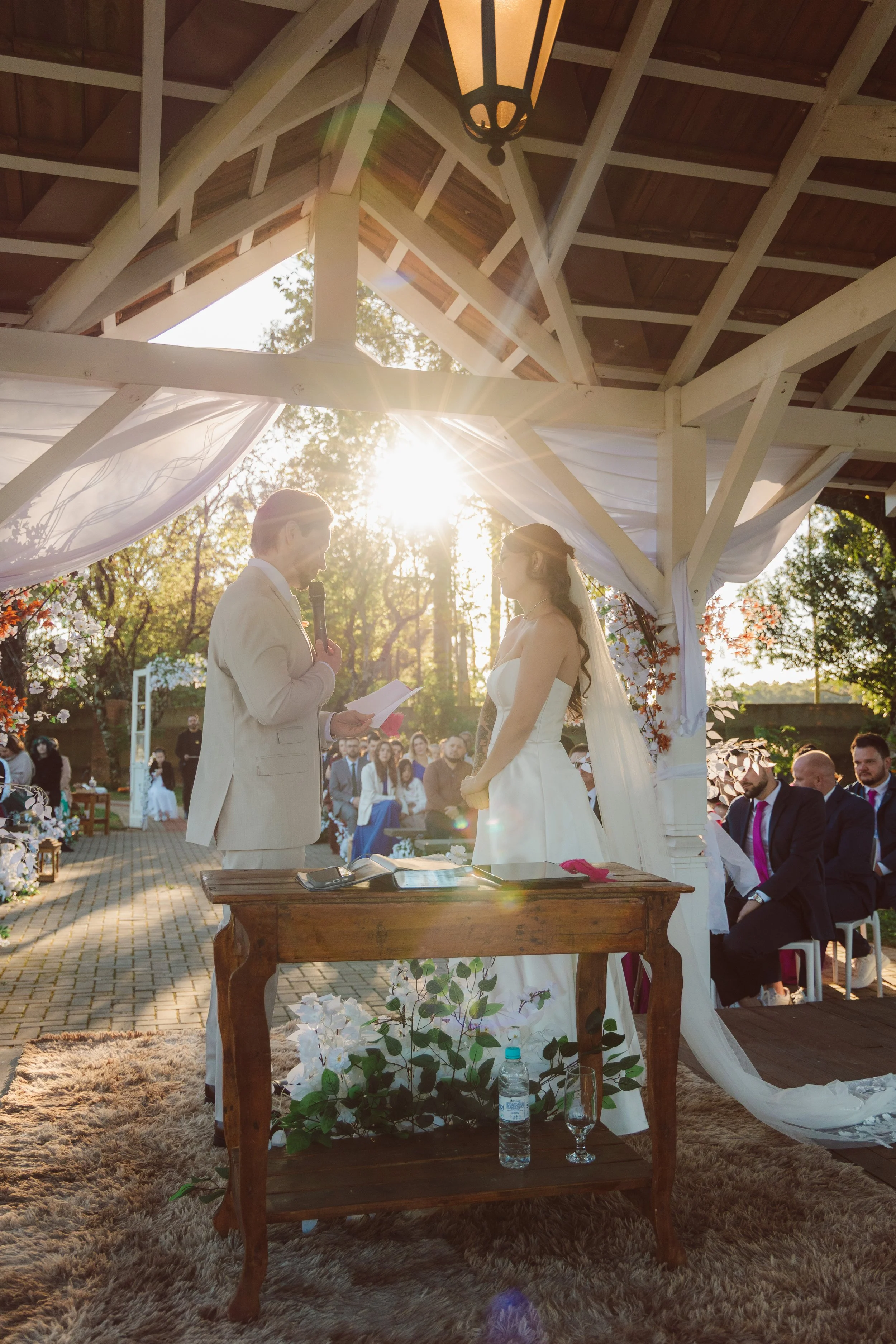 A wedding ceremony taking place outdoors under a wooden pavilion with sunlight shining through. The bride and groom are standing facing each other, with the officiant holding a microphone and paper, likely reading vows. Guests are seated in the backg