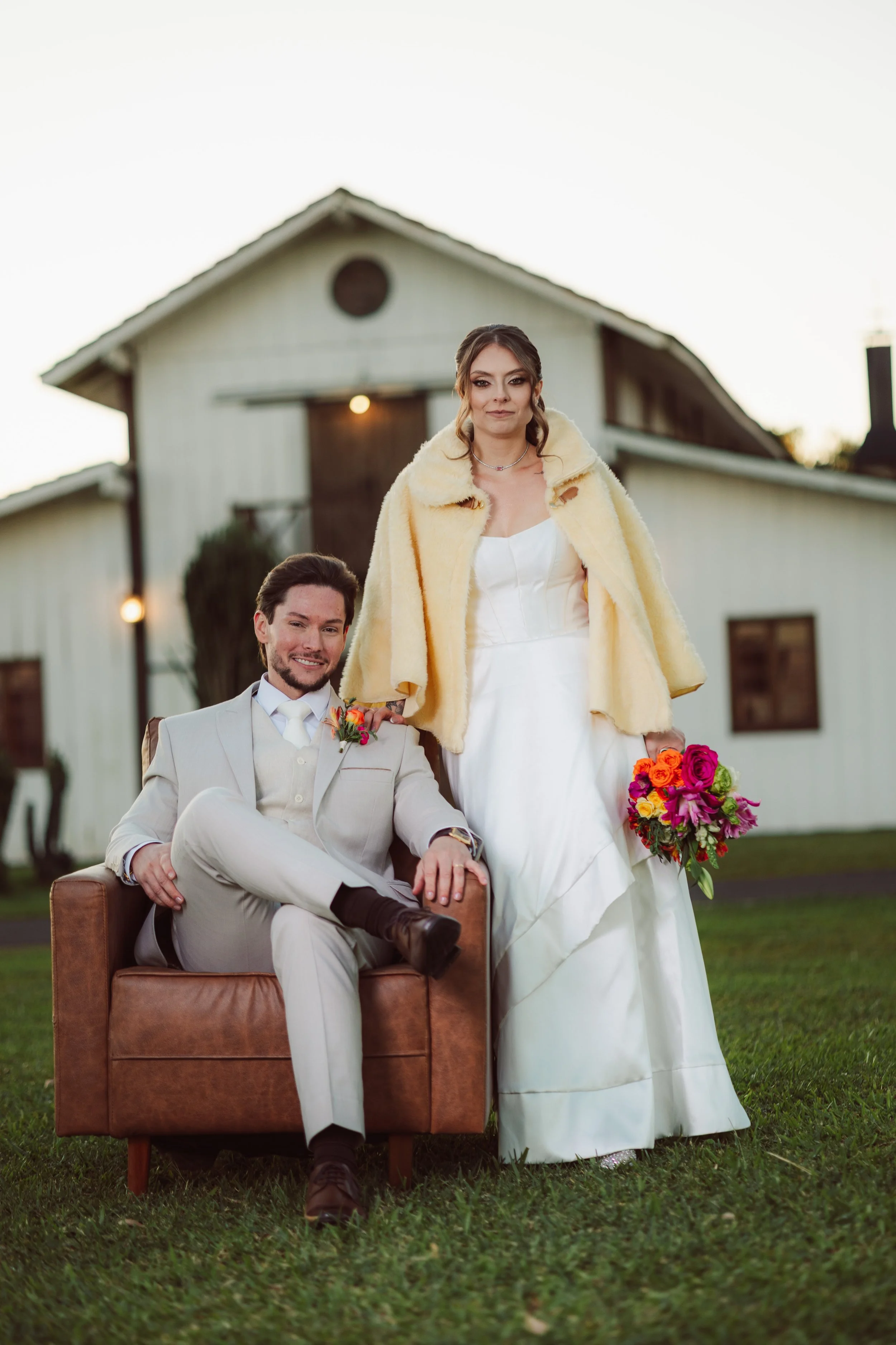 A bride in a white wedding dress with a yellow fur coat holding a bouquet of colorful flowers, standing next to a seated man in a light gray suit with a boutonniere, in front of a white barn during sunset.