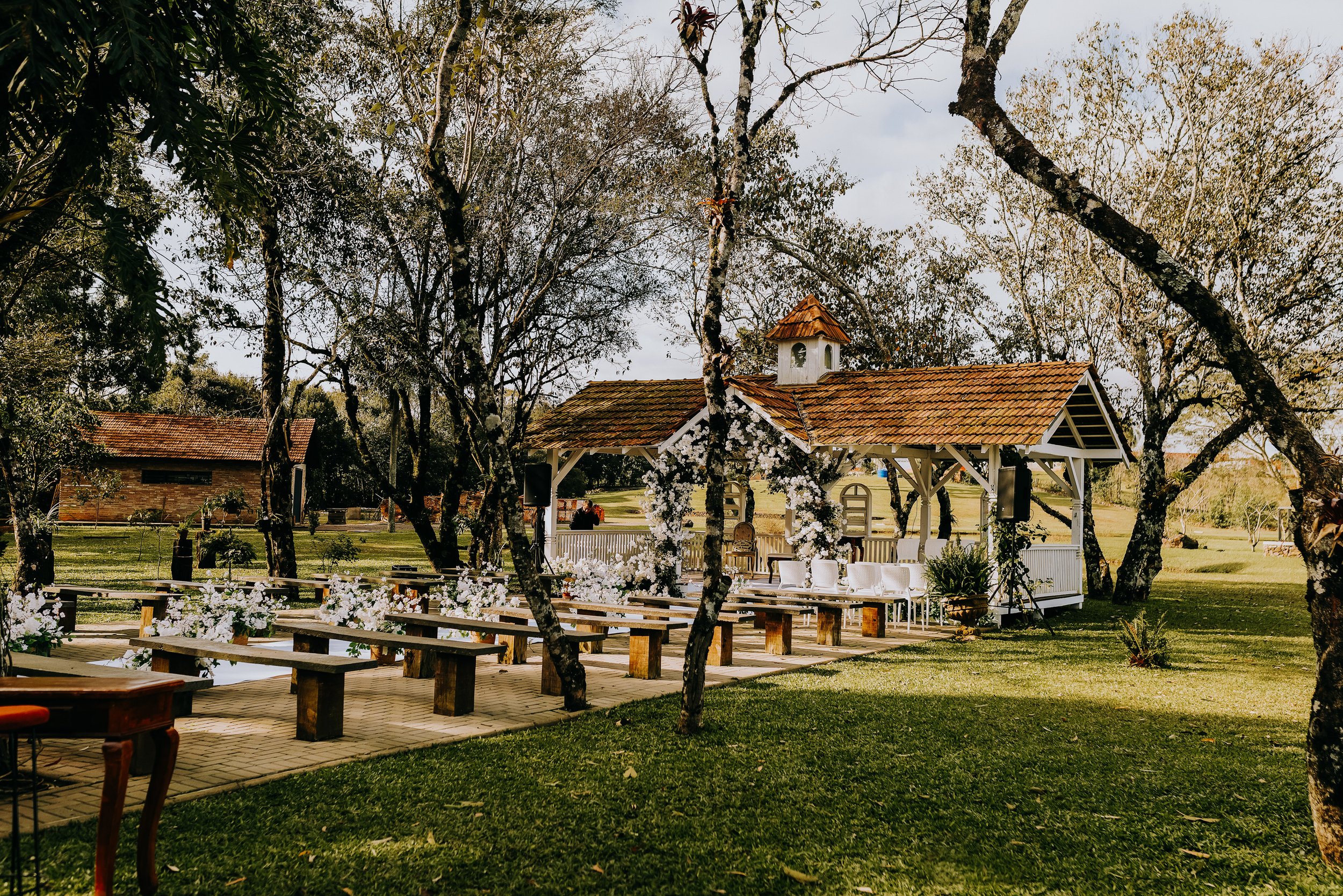 An outdoor wedding ceremony setup with a white gazebo decorated with white flowers, surrounded by trees, with chairs and benches arranged for guests.