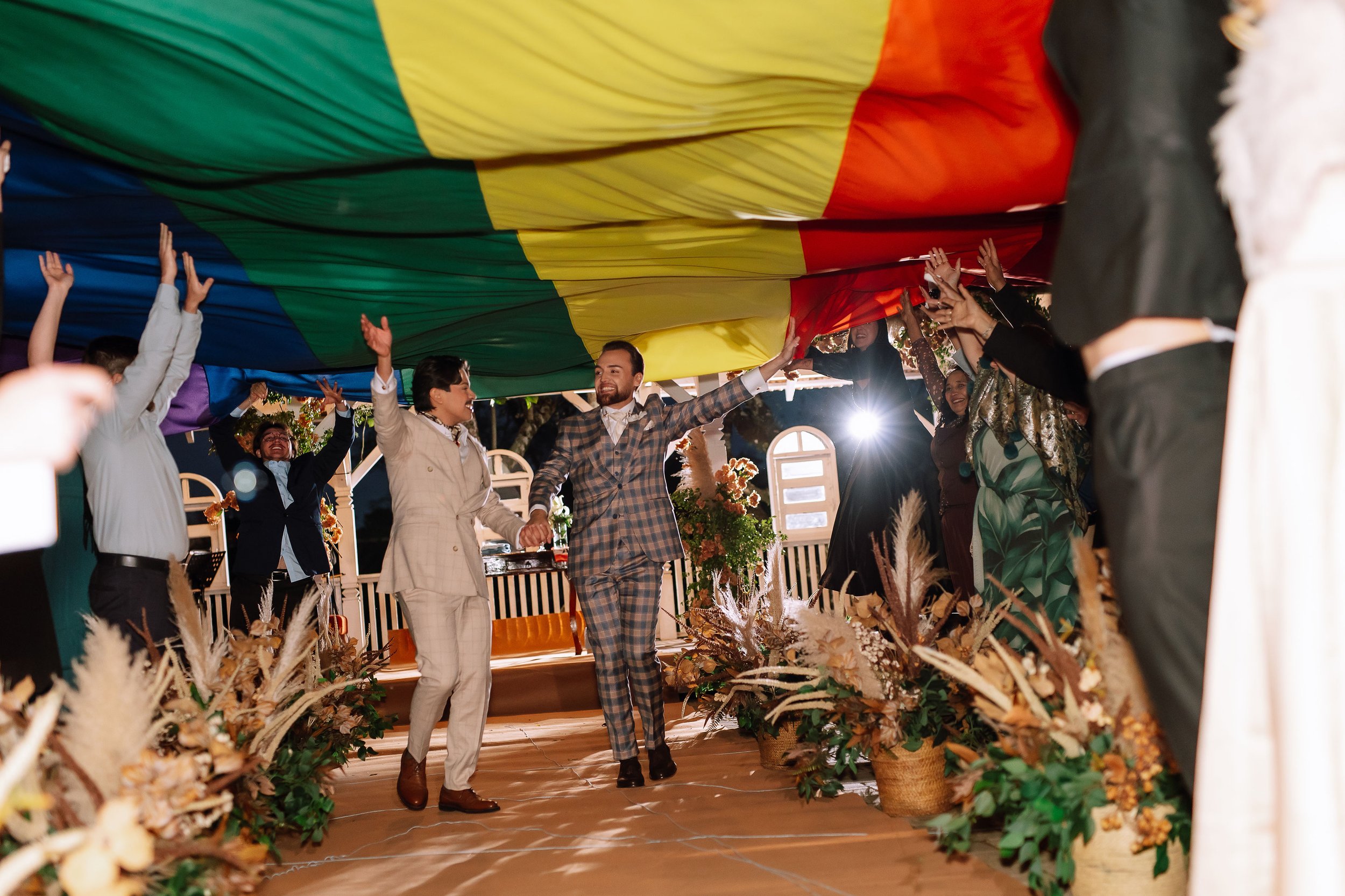 Two men are dancing under a large rainbow flag, with several people surrounding them. The setting appears to be a celebration or event, with floral decorations and bright lighting.