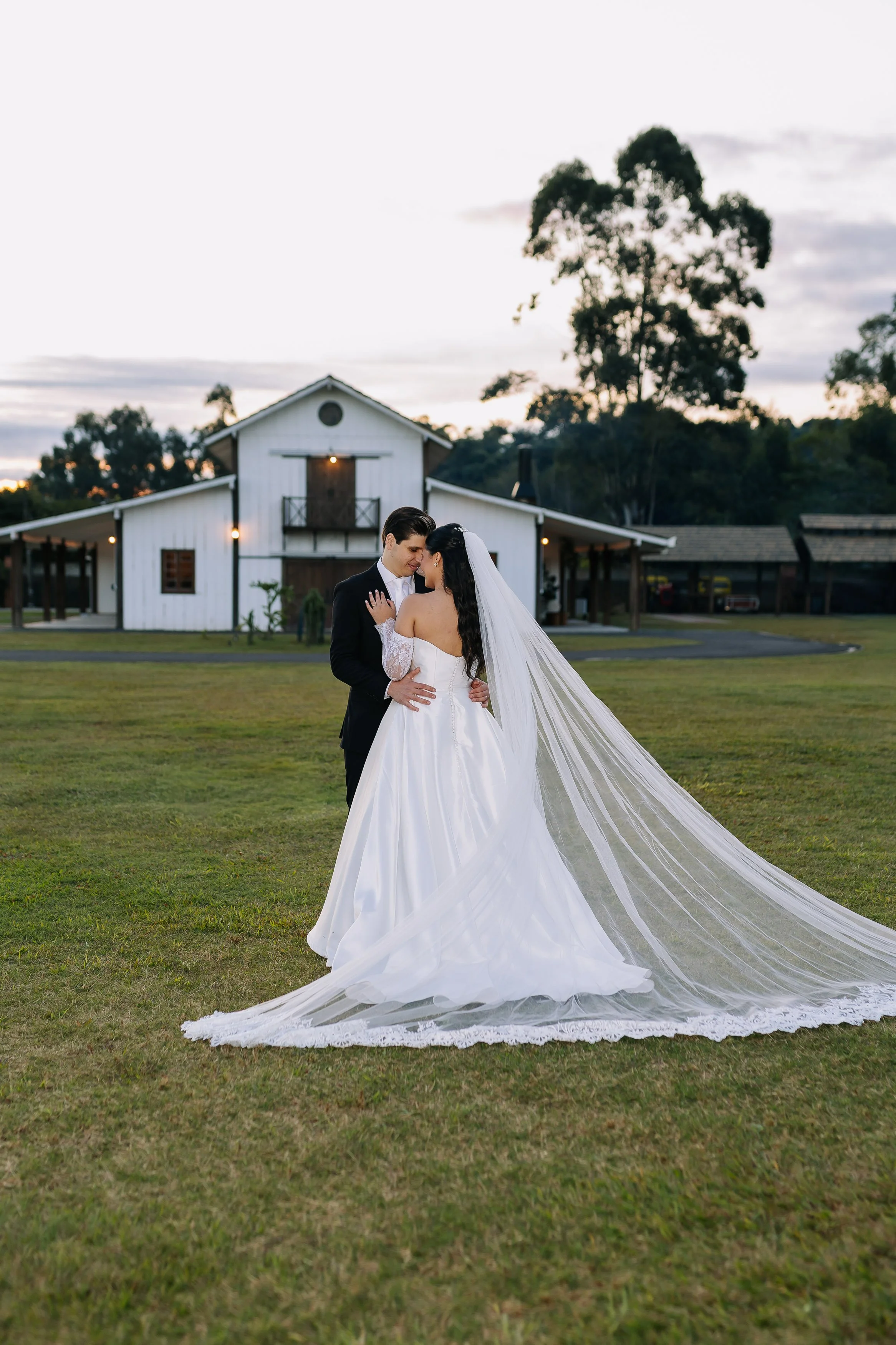 A bride and groom in wedding attire embrace outside a rustic white barn during sunset, with the bride's long train flowing on the ground.
