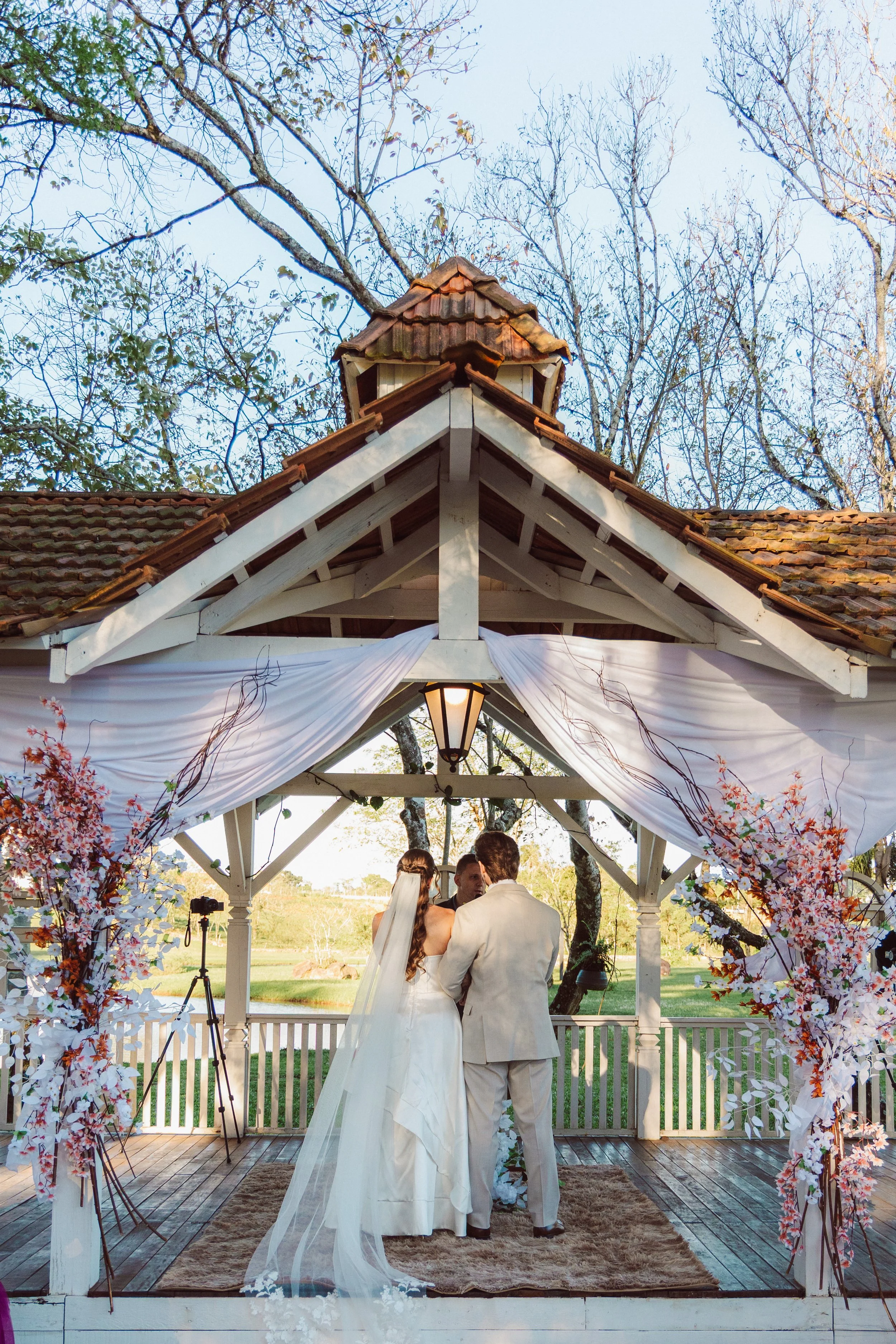 A bride and groom are standing together in a wedding ceremony under a decorated outdoor pavilion. The pavilion has white drapes and pink floral arrangements, with a small wooden roof and lantern hanging in the center. They are facing away from the ca