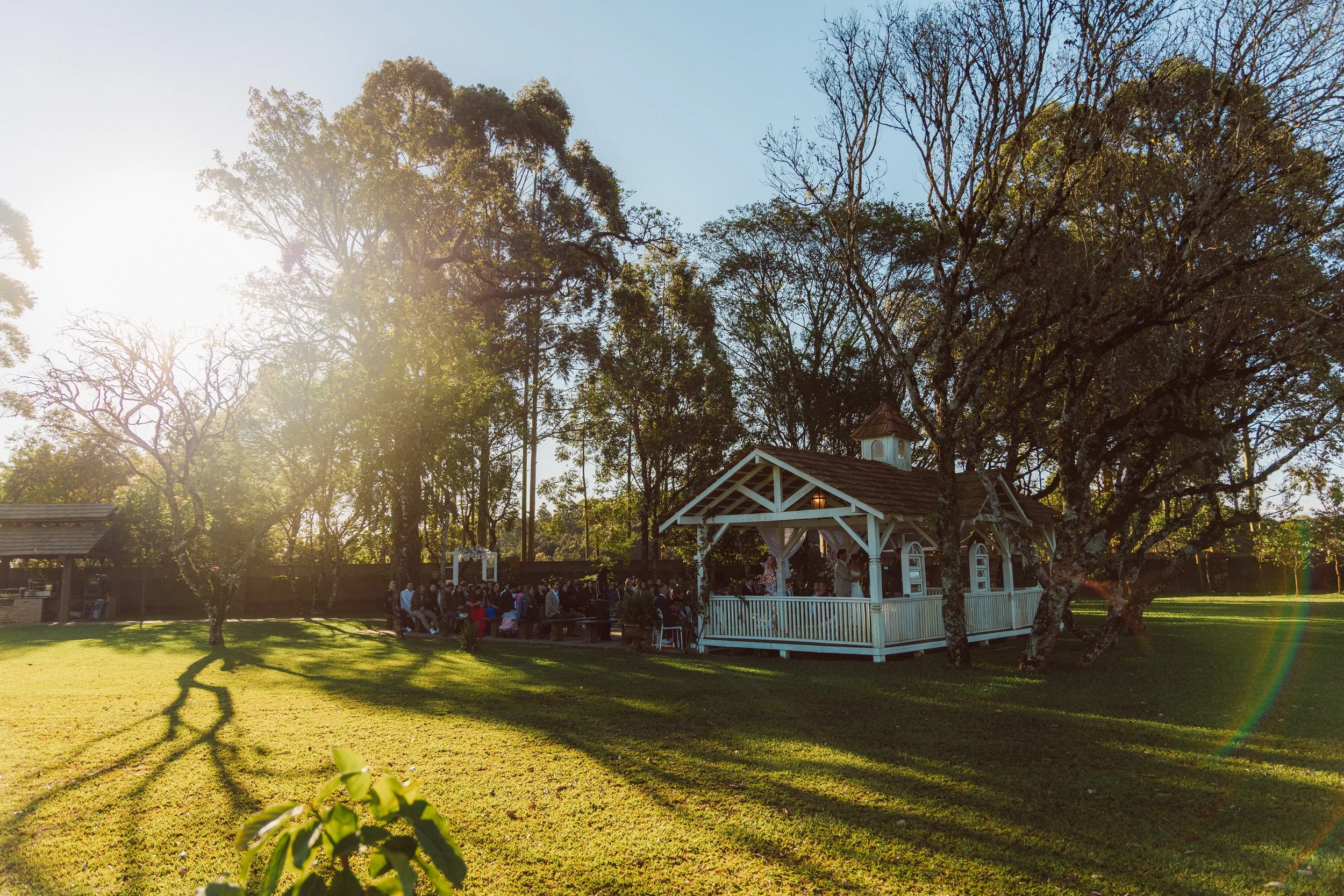 Outdoor wedding ceremony in a park with a white gazebo, trees, and guests seated on benches.