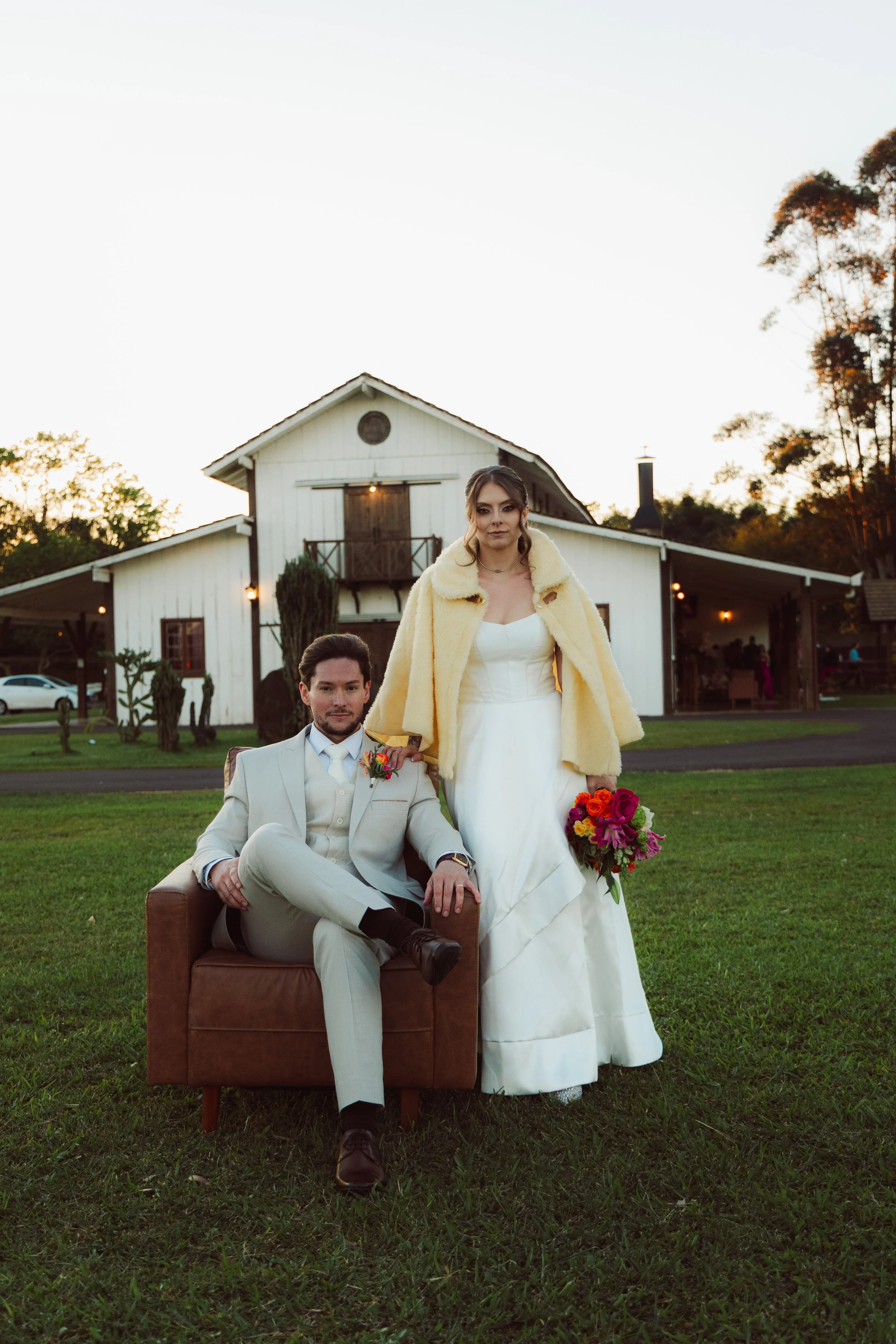 A bride and groom posing outdoors during sunset, with the bride standing and the groom sitting on a chair on a grassy lawn, in front of a white barn-style building.