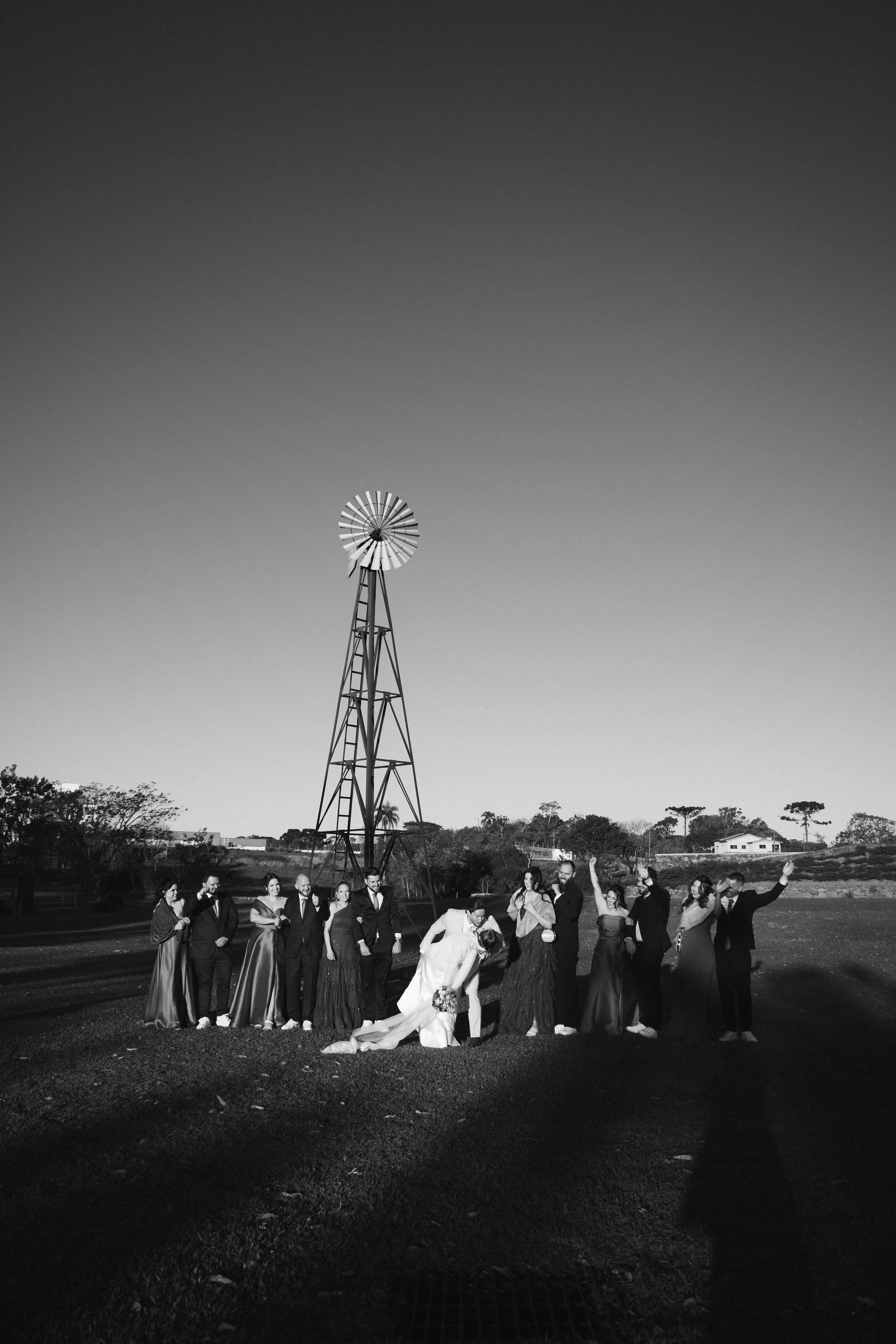 A group of people dressed in formal attire standing on a grassy field with a windmill in the background. The photo is in black and white, and the group appears to be celebrating or at a special event.