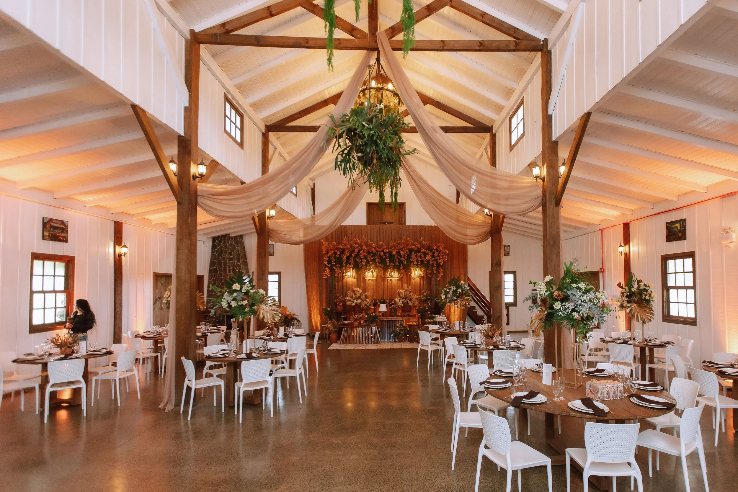 Event space with high vaulted ceiling, decorated with draped fabric, hanging greenery, and chandelier. Round tables with white chairs set for a celebration, floral arrangements, and nighttime lighting.