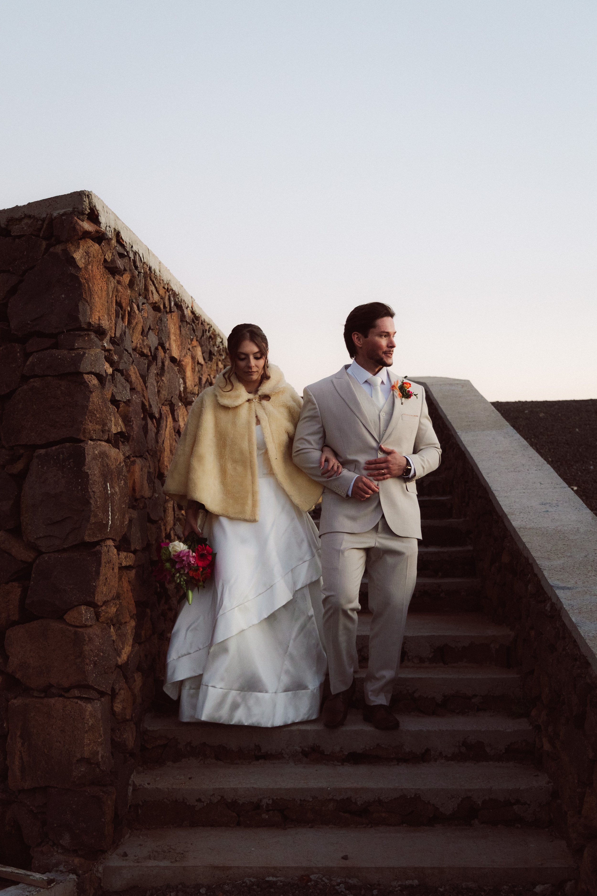 A bride and groom walking down the stairs outdoors during their wedding, with a stone wall on the left and a clear sky above.