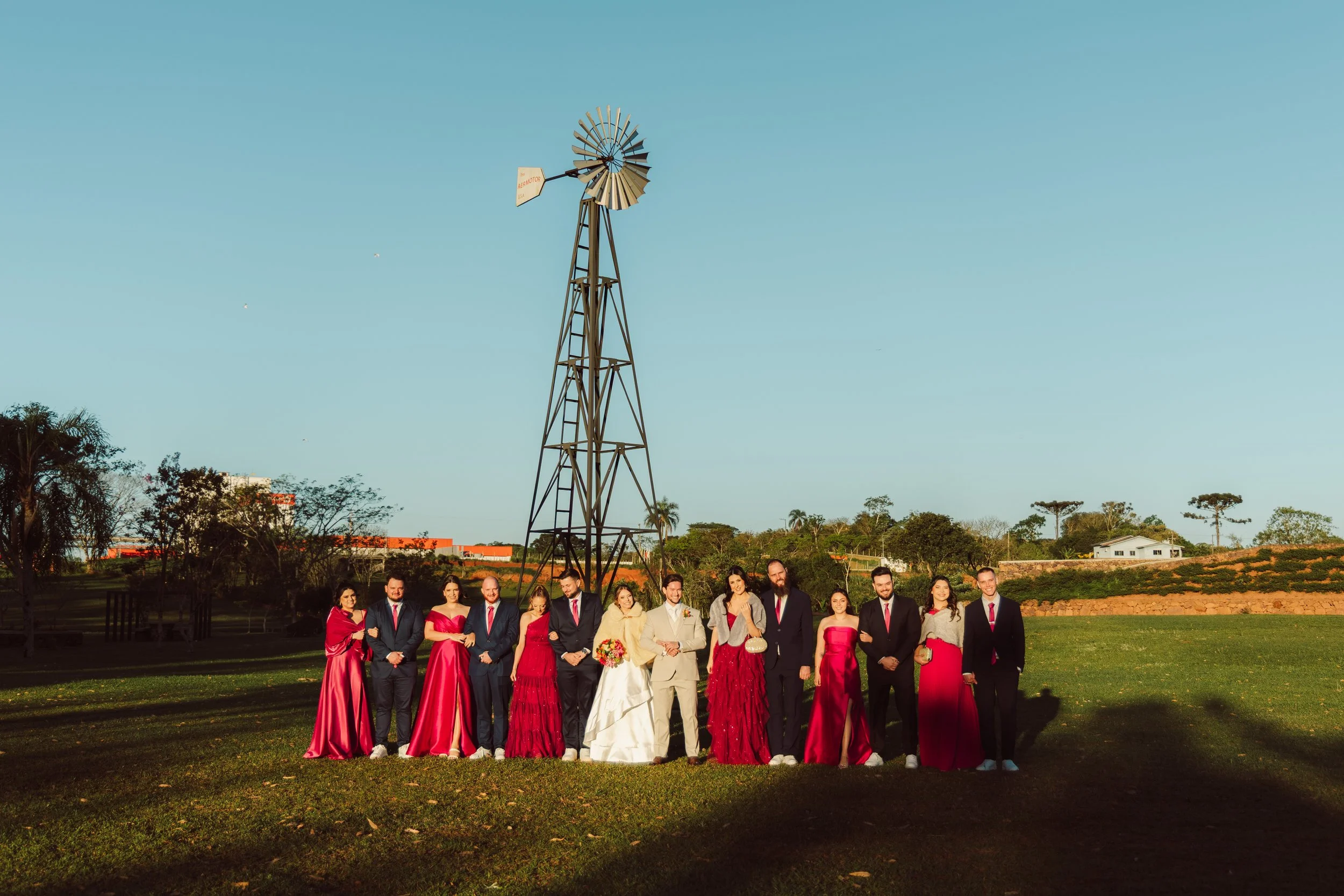 A group of people dressed in formal attire, including wedding dress and suits, standing outdoors on grass with trees, houses, and a windmill in the background on a clear day.