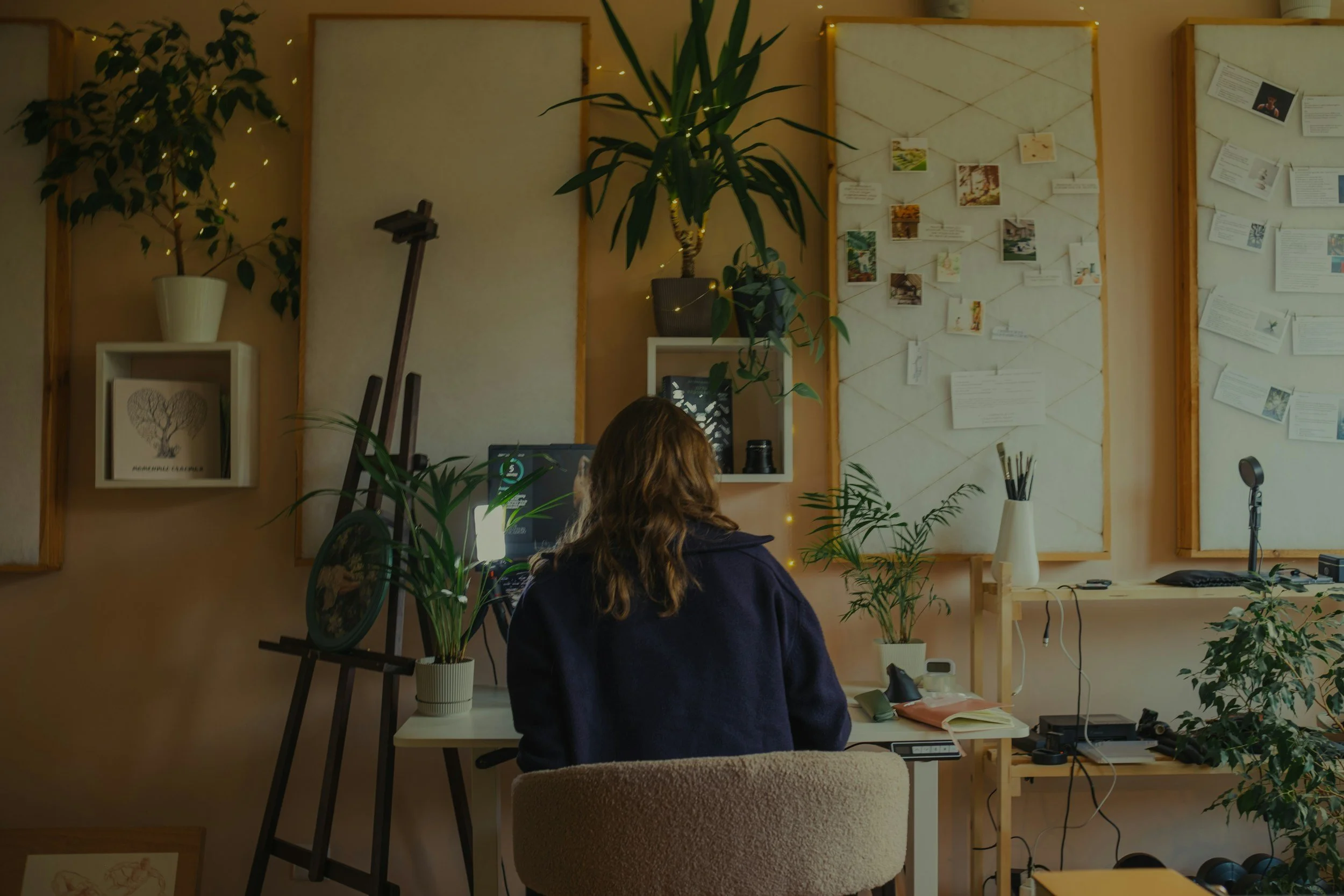 A woman with brown hair, wearing a dark blue sweater, is sitting at a desk in a cozy room decorated with potted plants, framed artwork, and photo displays on the wall. There is a computer monitor, keyboard, and various office supplies on the desk.