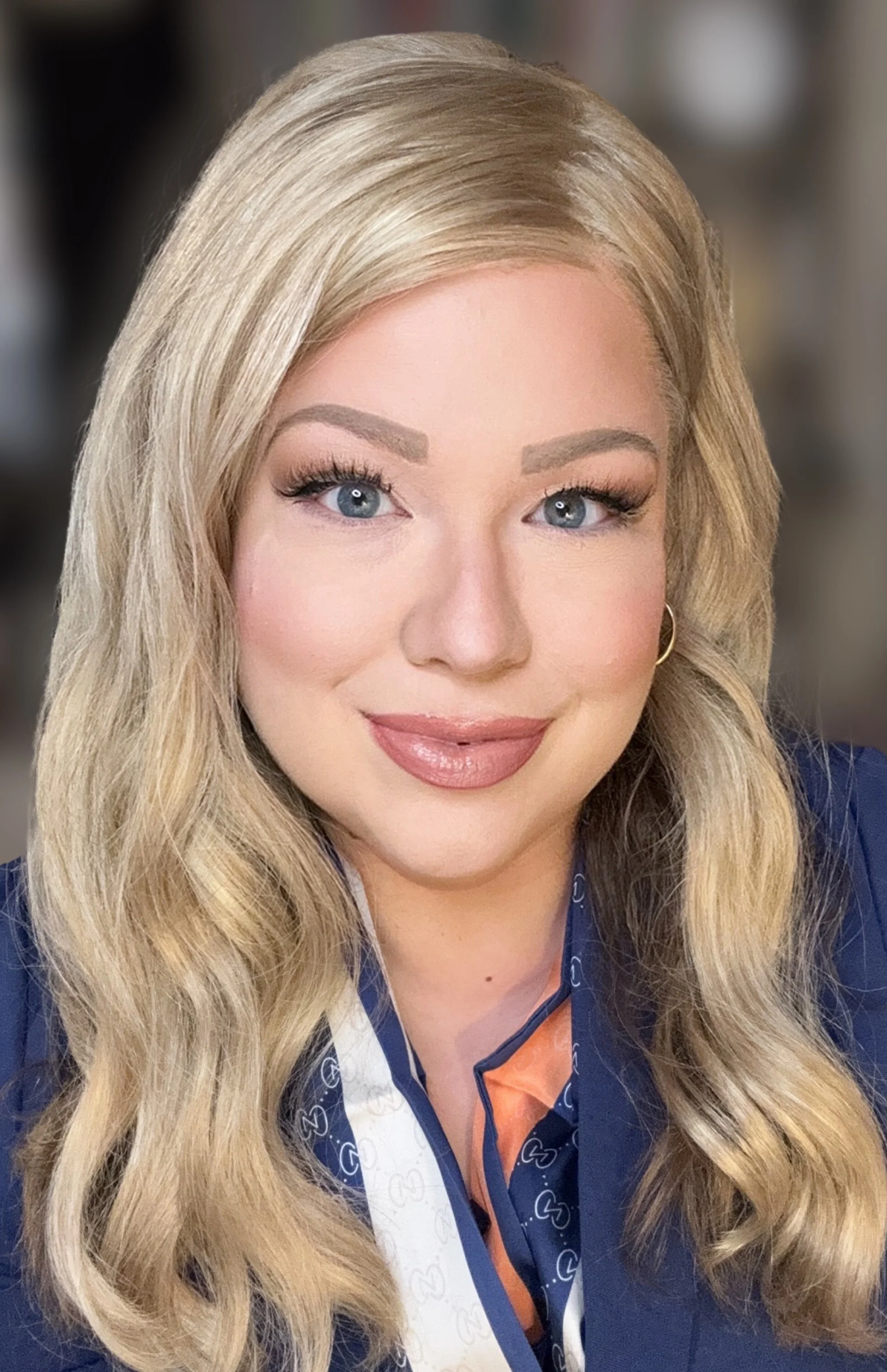 A woman with long blonde hair, blue eyes, and makeup, wearing a navy blue jacket and a white sash with blue heart patterns, smiling for the camera.