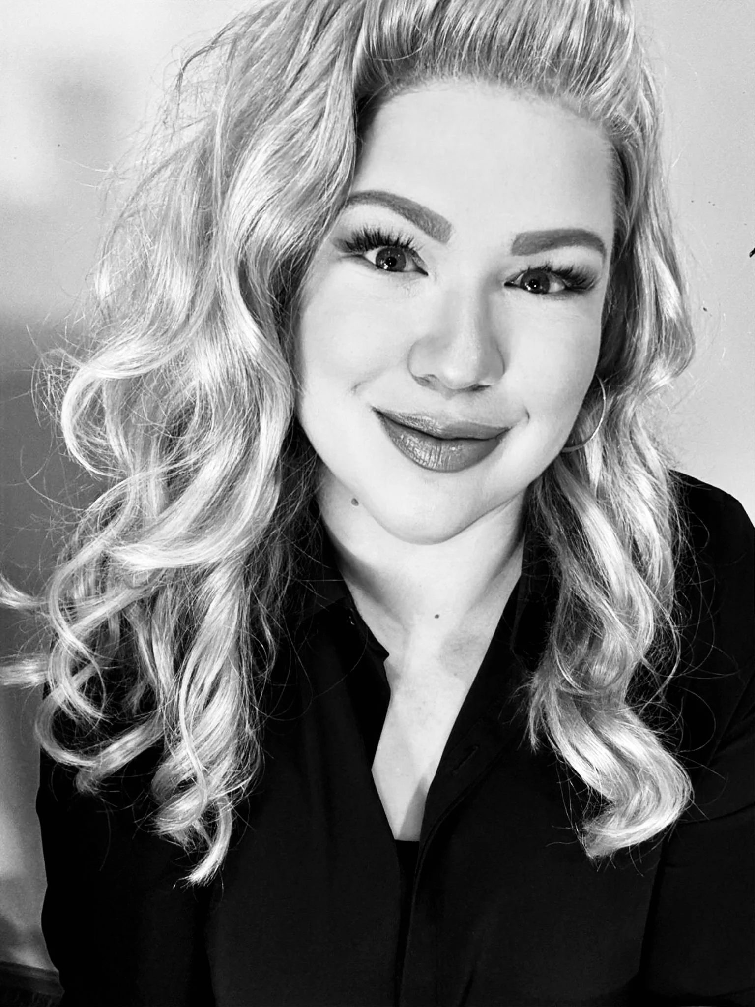Black and white portrait of a woman with long, wavy hair. She is smiling slightly, wearing makeup including eyeshadow, eyeliner, and lipstick, and dressed in a dark top.