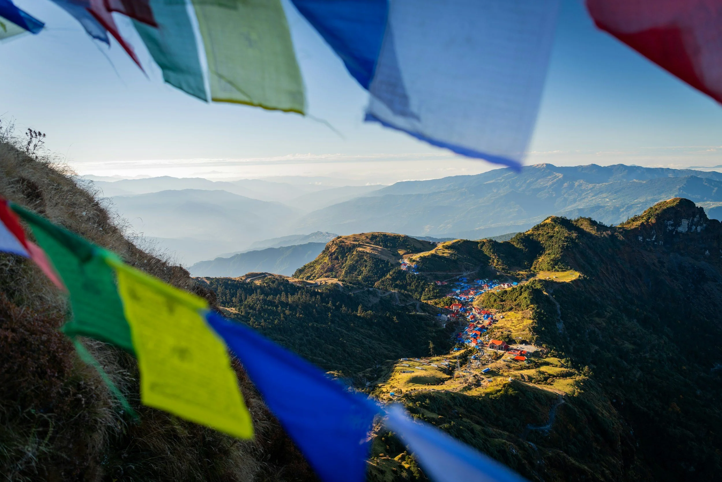 Vistas de montañas y un valle con casas, con banderas de plegarias de colores en primer plano y una vista panorámica del cielo y las montañas en el fondo.