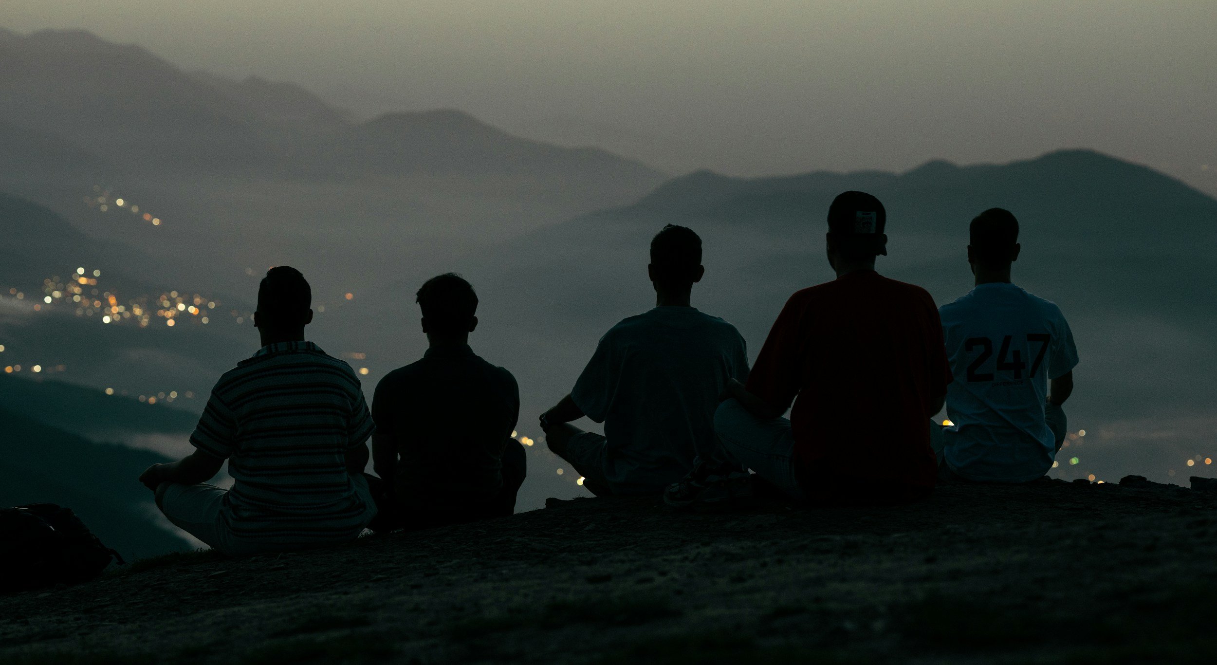 Cinco personas sentadas en una colina observando el paisaje montañoso y las luces de una ciudad en la distancia durante el atardecer o anochecer.