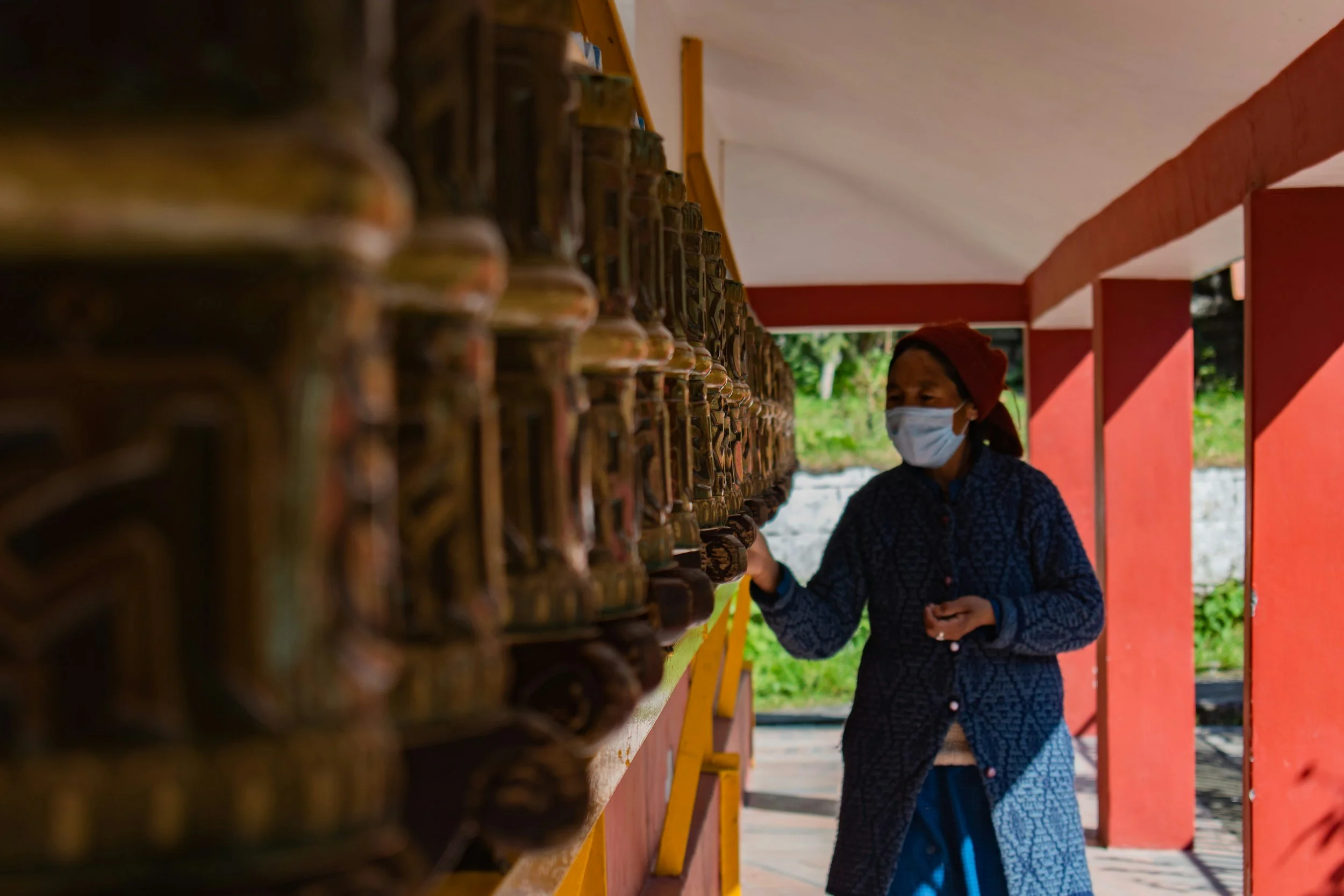 Mujer con mascarilla observando objetos o figuras en una línea en un espacio abierto con paredes naranjas y fondo natural verde.