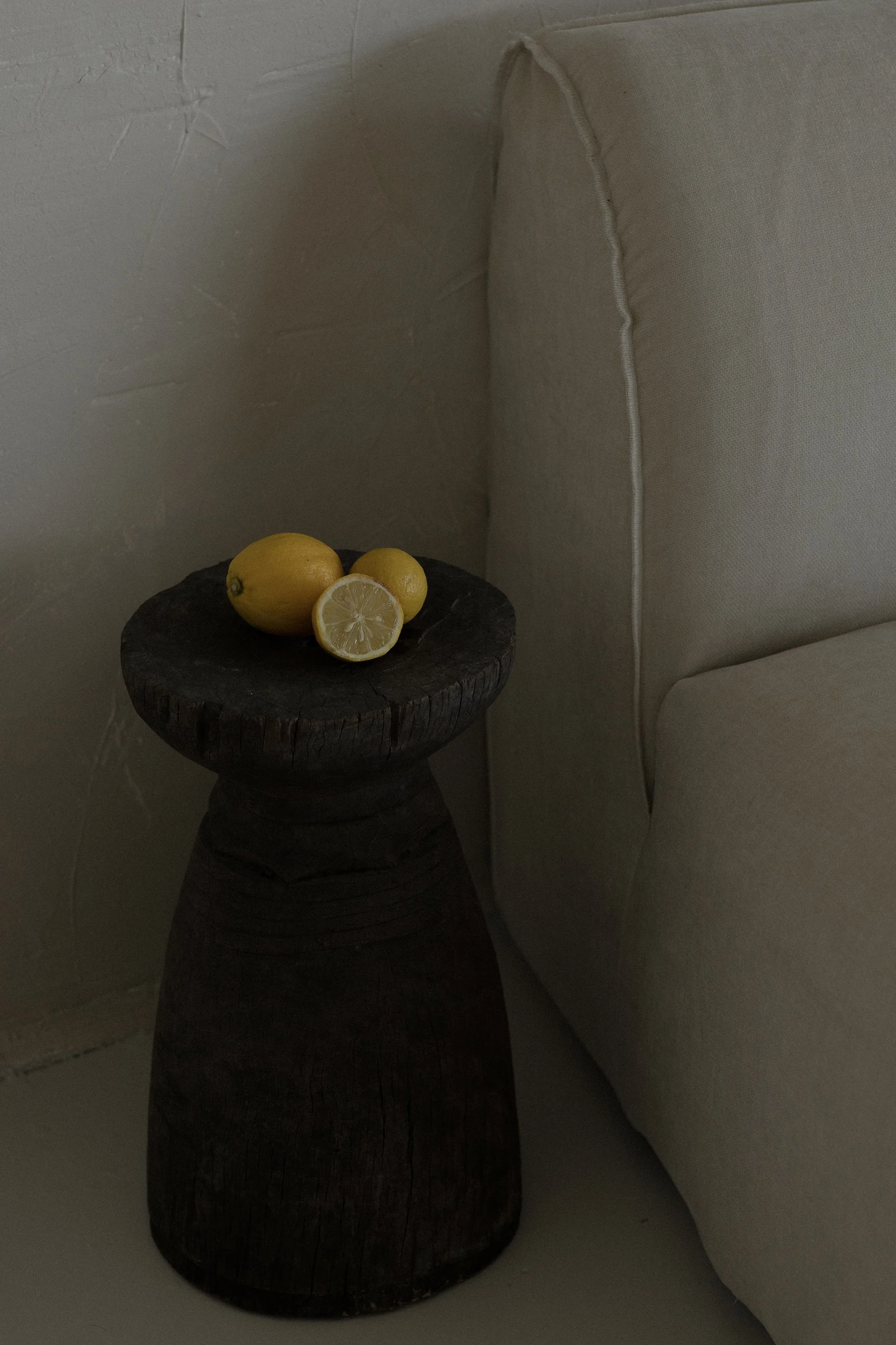 A dark wooden stool with a lemon, a cut lemon, and a small lemon on top of it. A beige wall with textured paint and a corner of a white couch with a fabric cover.