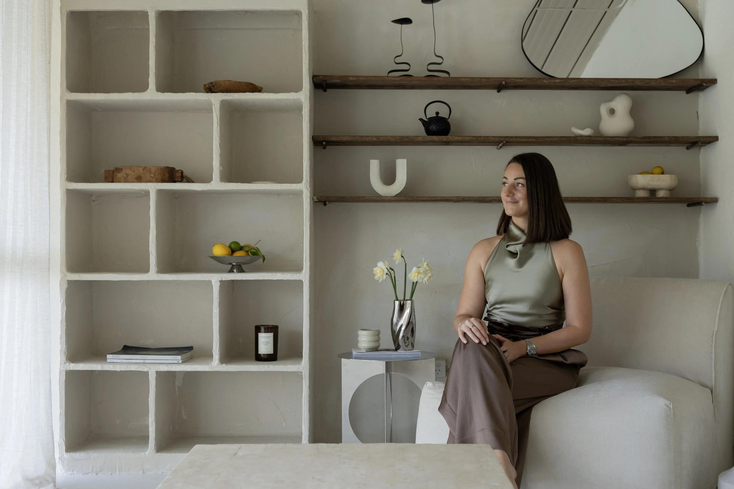 A woman sitting on a beige sofa in a minimalist room with shelves and decorative objects, including a black teapot, a serif letter U, a white abstract sculpture, a mirror, and a bowl with lemons and limes.