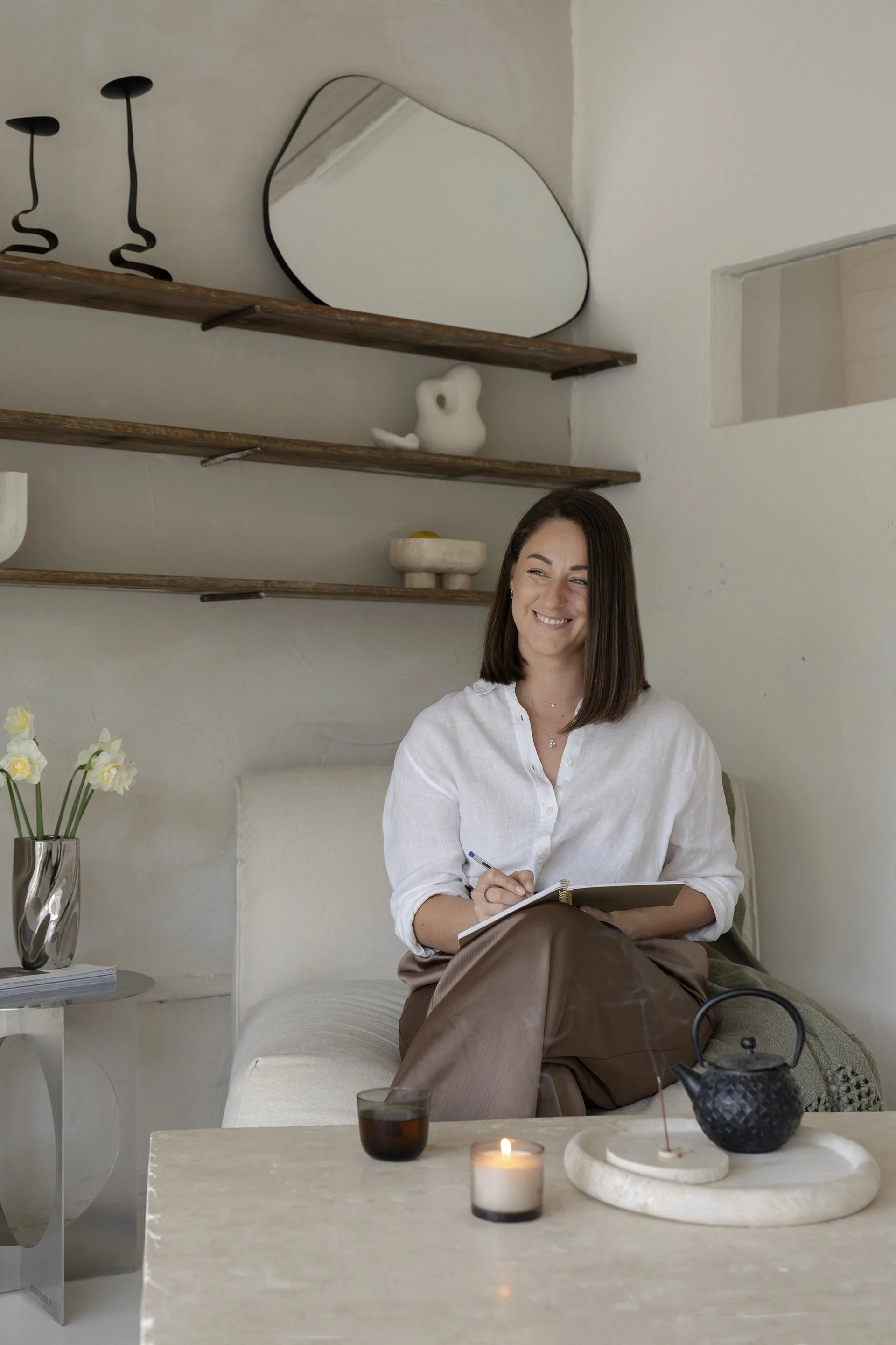 A woman sitting on a beige sofa in a bright, minimalist living room, smiling while writing in a notebook. The room has a side table with a vase of white flowers, candles, and decorative objects. Wooden shelves with art pieces are on the wall behind her.