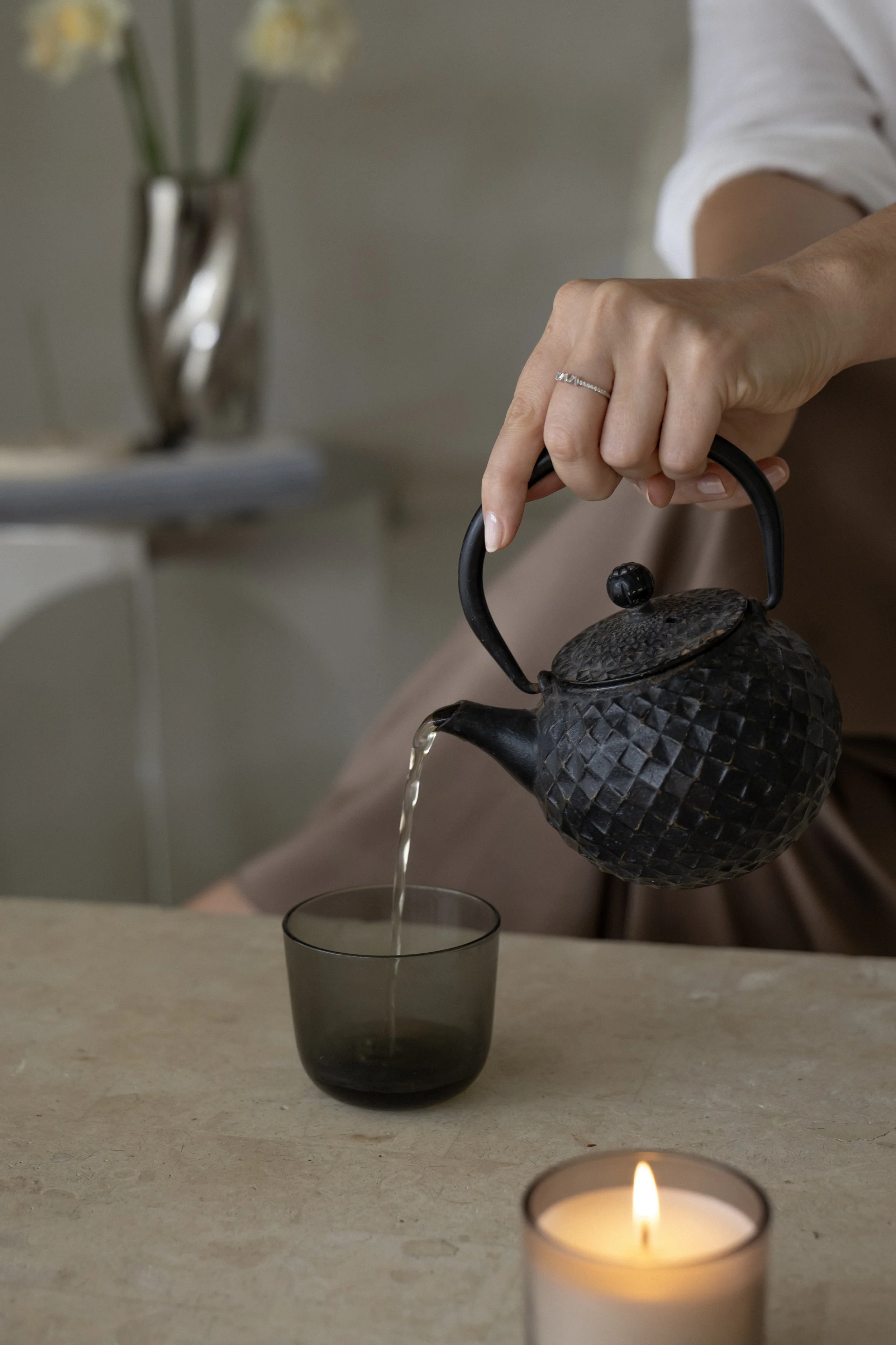 A person is pouring tea from a textured black teapot into a clear glass cup on a beige surface. A lit candle is in the foreground, and a vase with yellow flowers is blurred in the background.