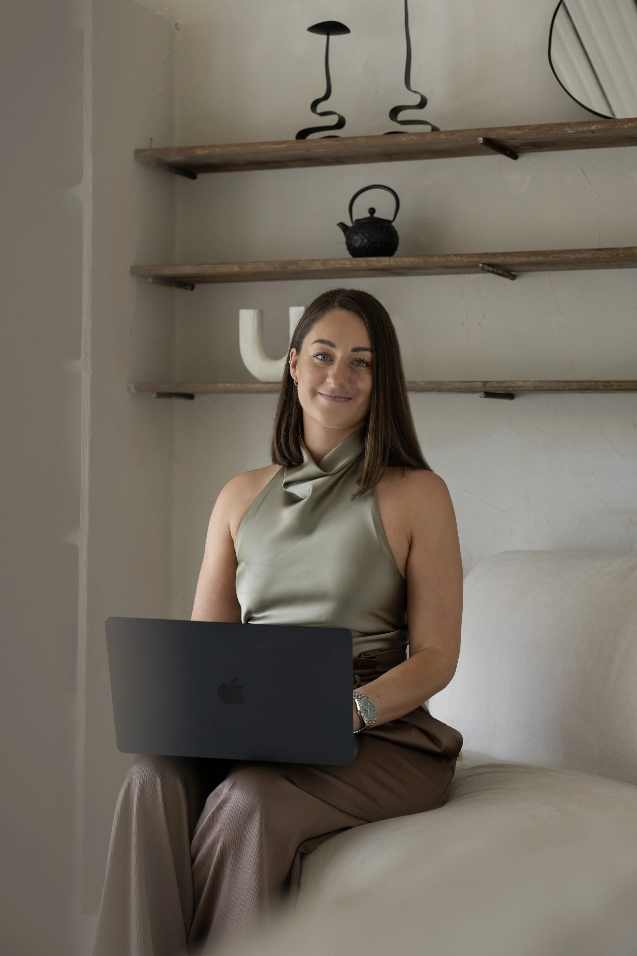 A woman with shoulder-length brown hair sitting on a bed with a laptop on her lap, wearing a sleeveless satin top and brown pants, in a room with wooden shelving and decorative items.