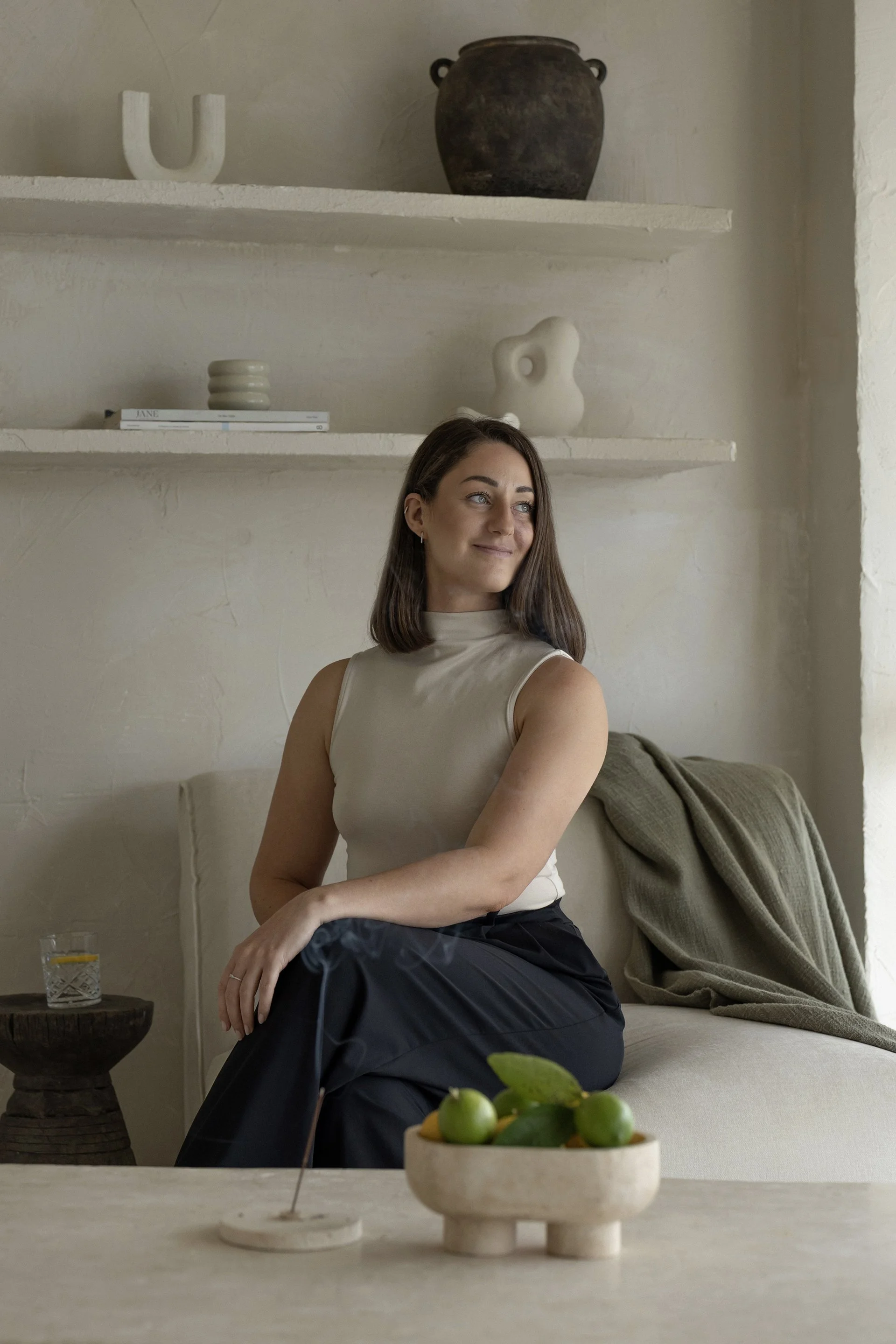 A woman sitting on a beige sofa in a room with minimalist decor, with a small wooden table in front, a bowl with green fruits, a glass of water, and a candle. She has shoulder-length brown hair, wears a sleeveless beige top, and looks to her left with a smile.