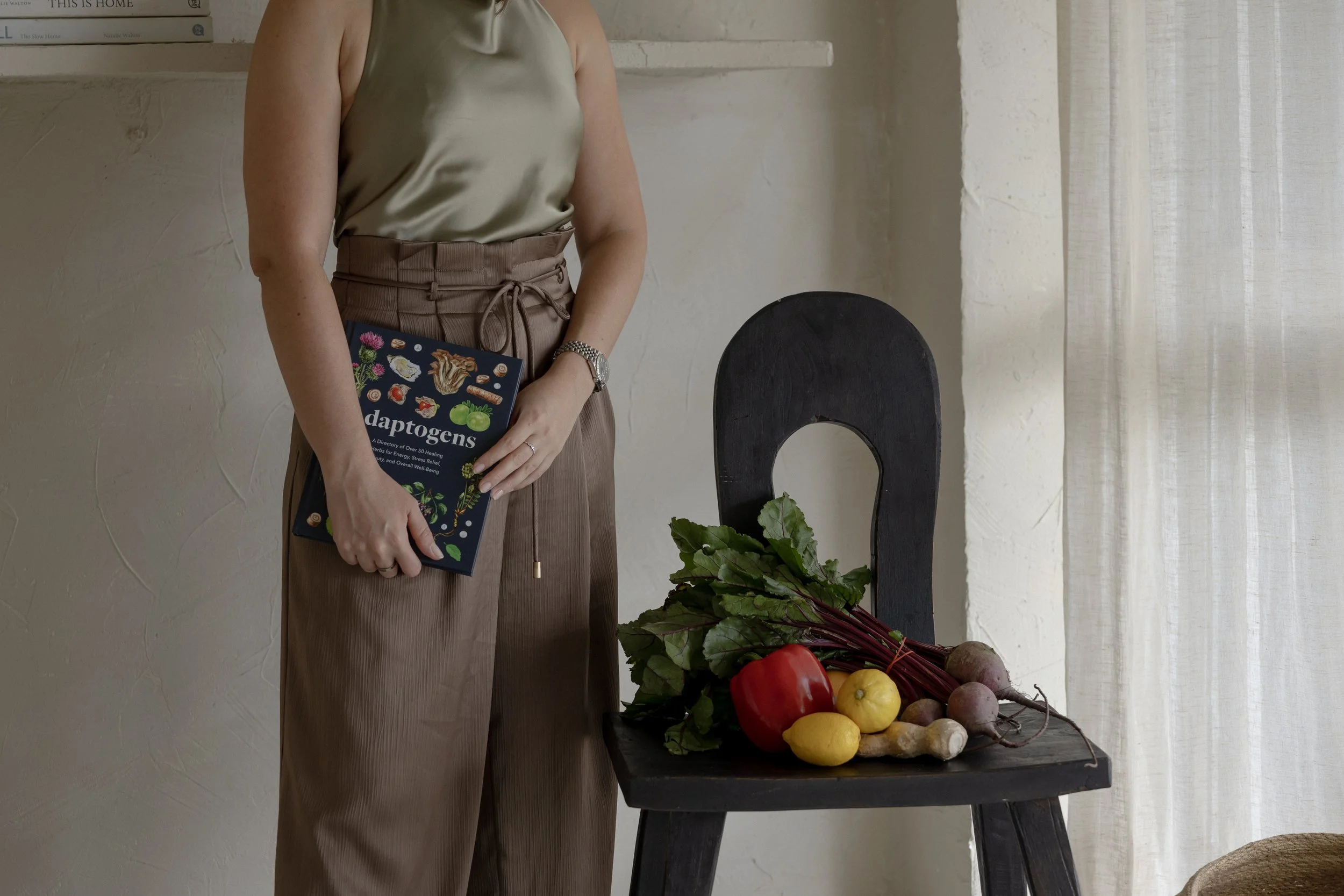 A woman holding a book titled 'Daptogens' standing next to a black table with fresh vegetables including beets, lemons, and red bell peppers, in a room with textured beige walls and a sheer curtain.