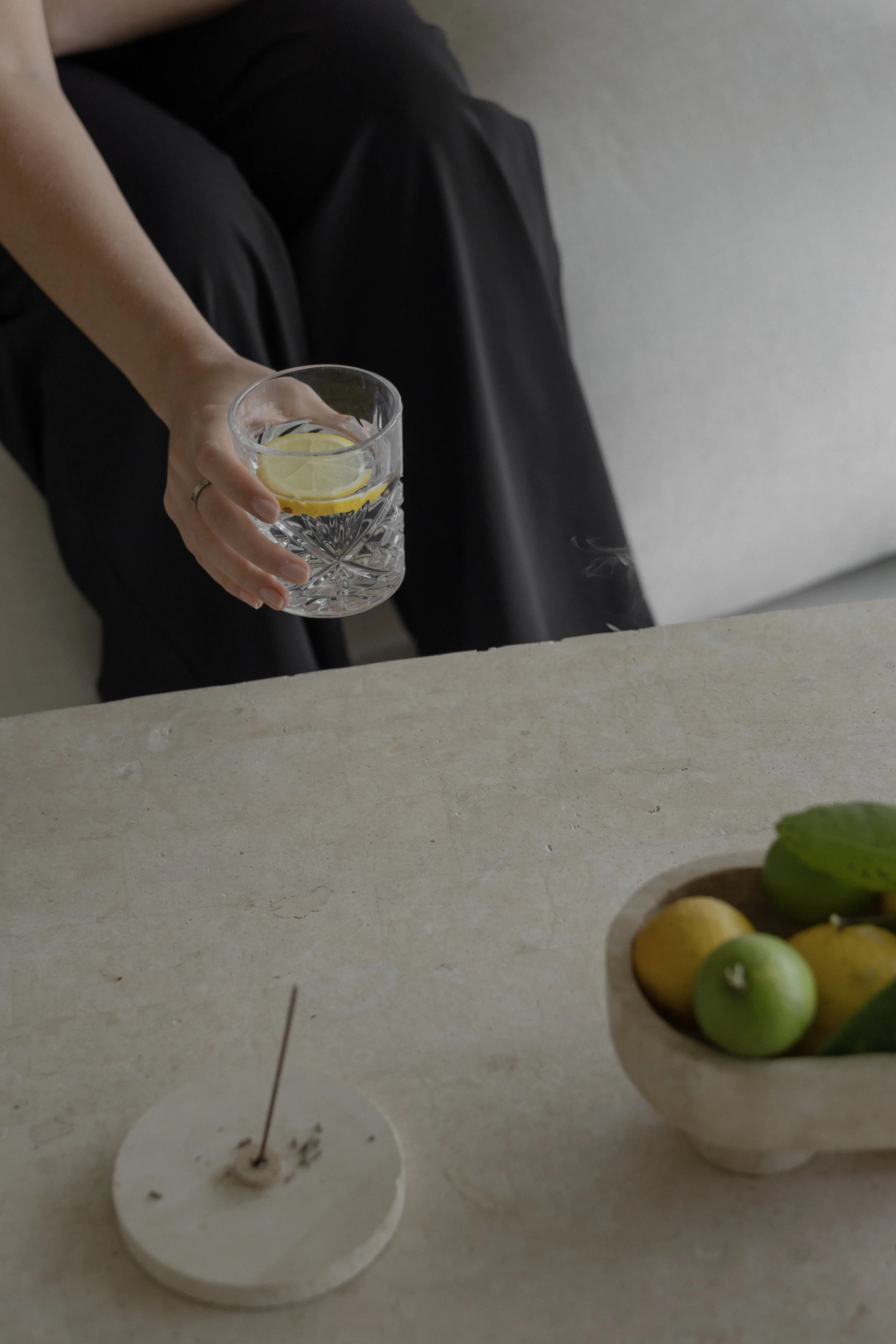 A person is holding a glass of water with lemon slices in it, sitting on a beige surface with a bowl of green apples and yellow lemons in the background.