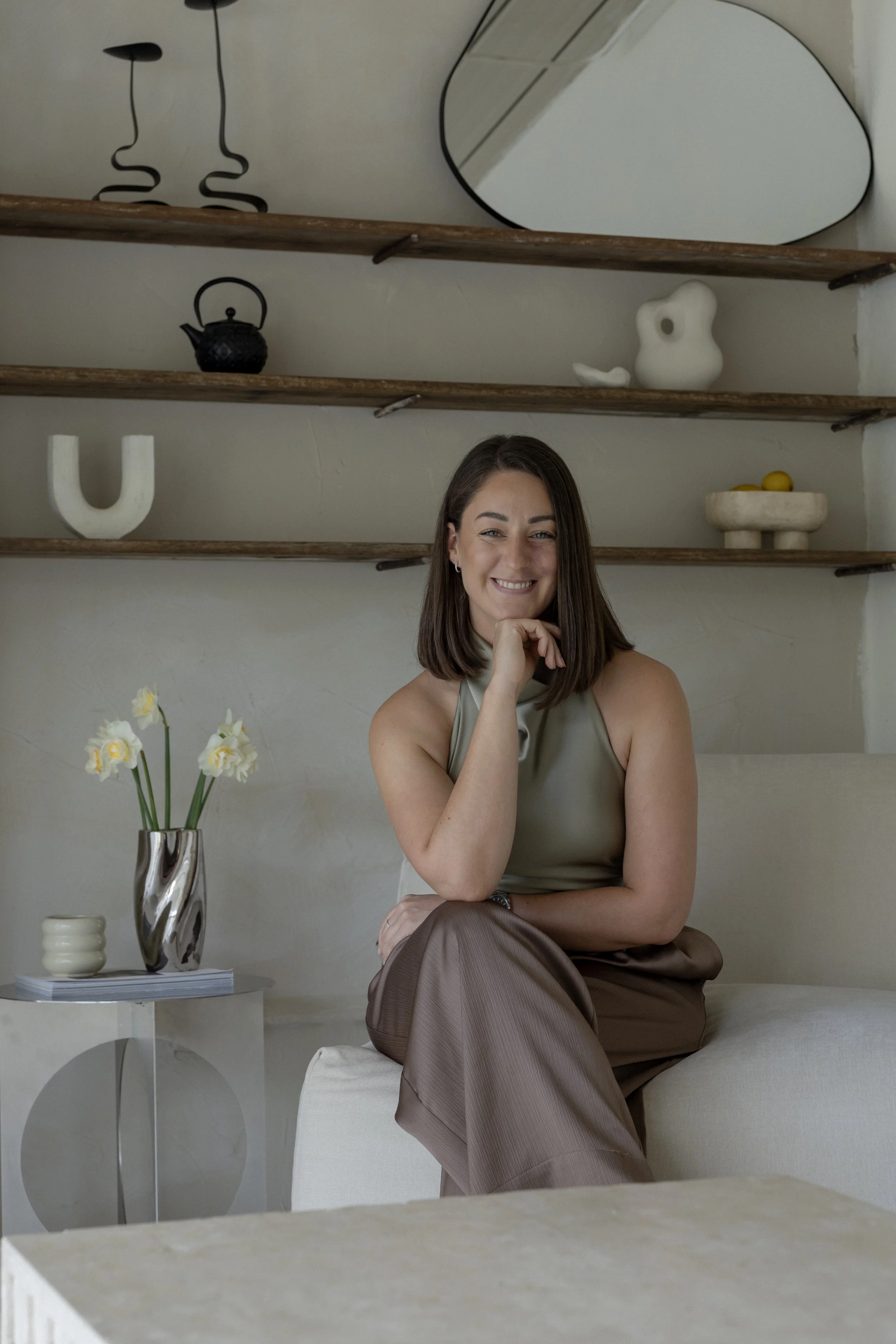 A woman with brown hair smiling, sitting on a beige couch, wearing a sleeveless beige top and brown trousers. Behind her, there are decorative shelves with minimalist art, a black teapot, a white abstract sculpture, and a bowl with yellow lemons. A vase with white flowers is on a side table next to her.