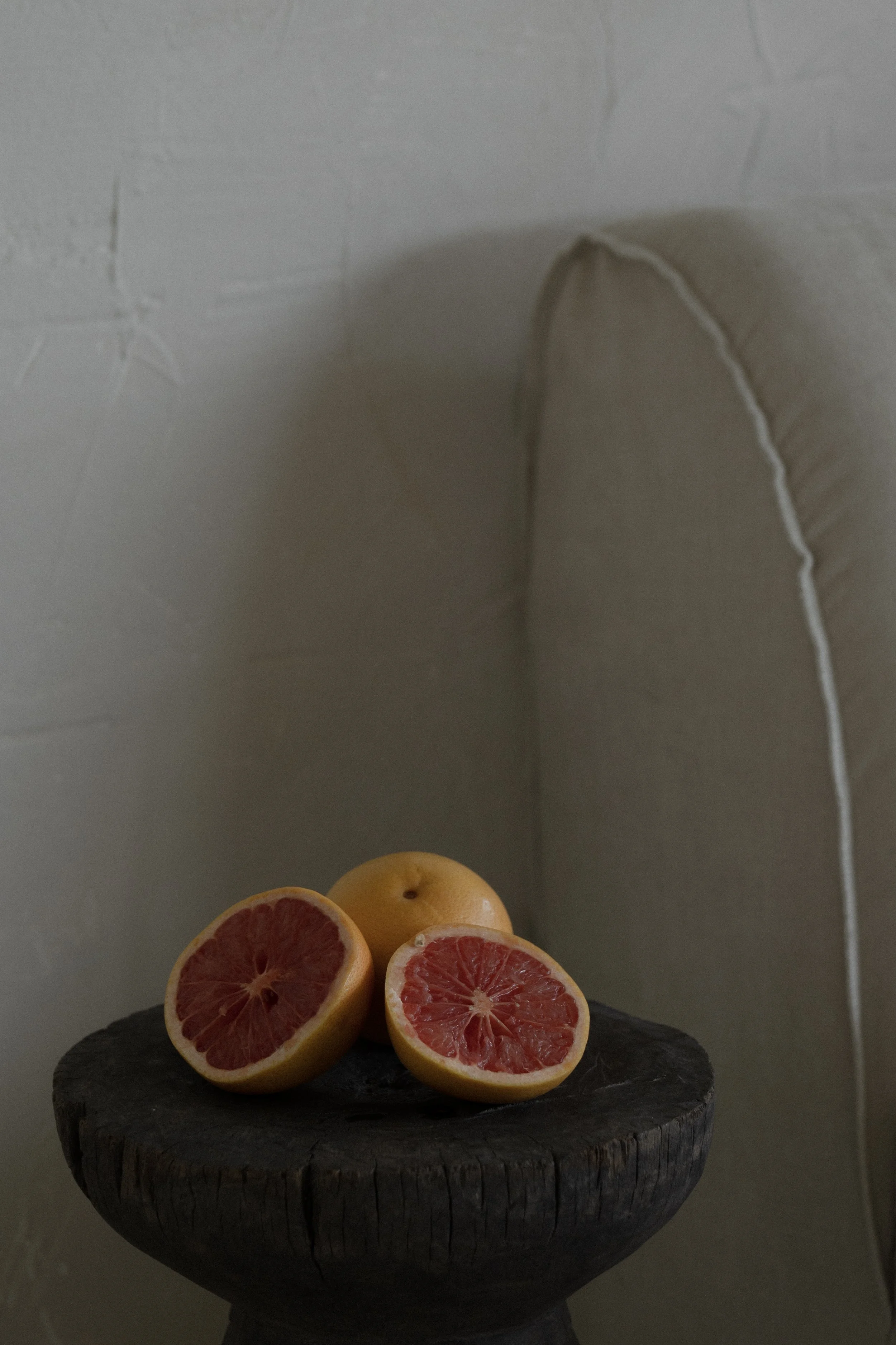 Several halved grapefruits on a dark stone surface, placed on a wooden stand with a textured gray background and visible wall seams.