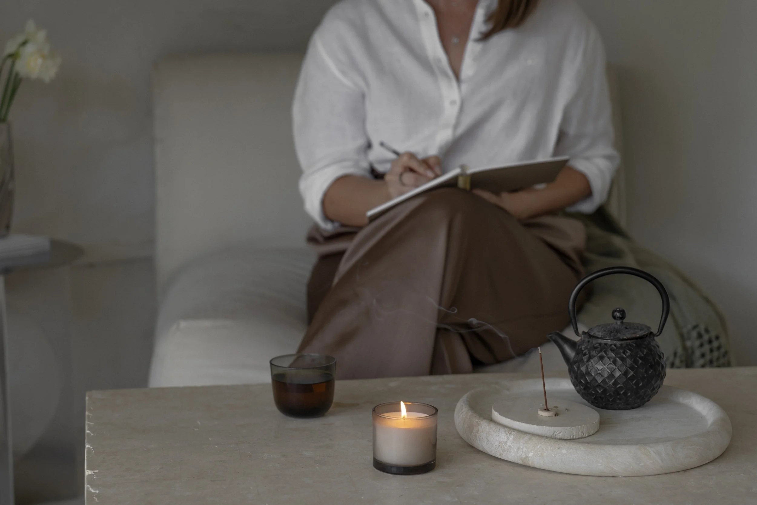 A woman in a white shirt is sitting on a white couch, holding a notebook and pen. On the table in front of her, there are lit candles, a black teapot, a glass of tea, and decorative items including a black textured vase and a white rock tray.