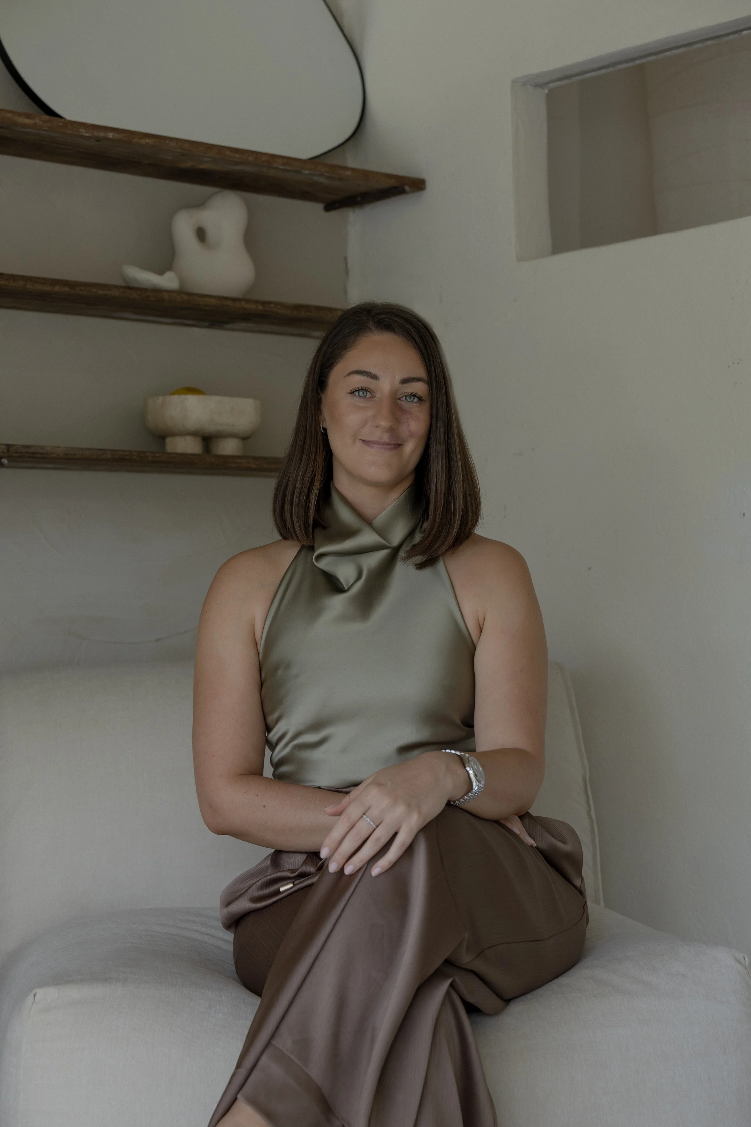 A woman with shoulder-length brown hair, wearing a shiny green sleeveless top, is sitting on a beige couch with her legs crossed. She has a watch on her left wrist and a ring on her right hand. Behind her, there are wooden shelves with decorative items, including a white abstract sculpture and a shallow bowl with yellow fruit. The room has neutral-colored walls and a small rectangular opening in the wall.