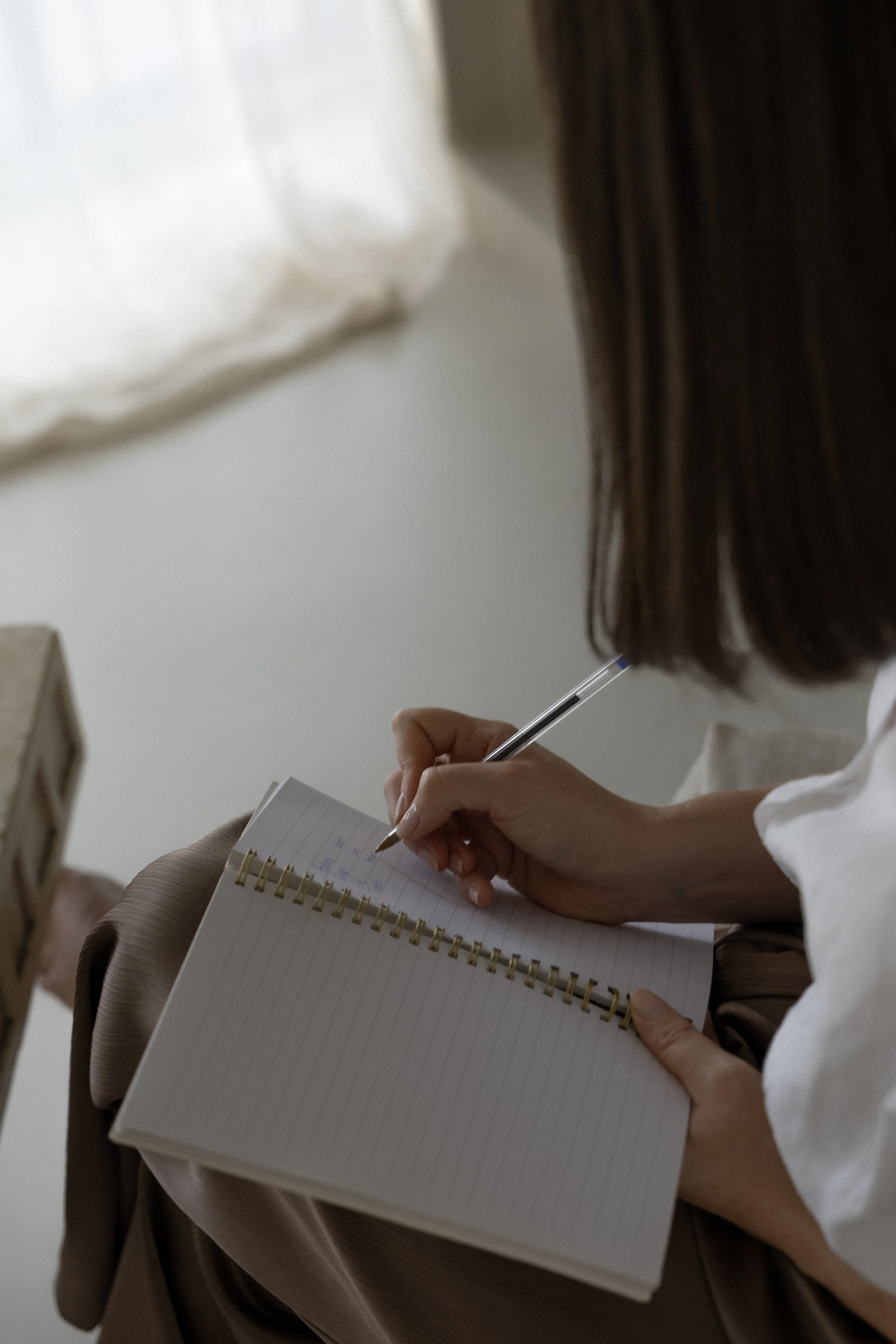 A person with long brown hair writing in a spiral notebook, with a white shirt and a tan coat or blanket draped over their lap. Another person's hand is visible holding the notebook steady.