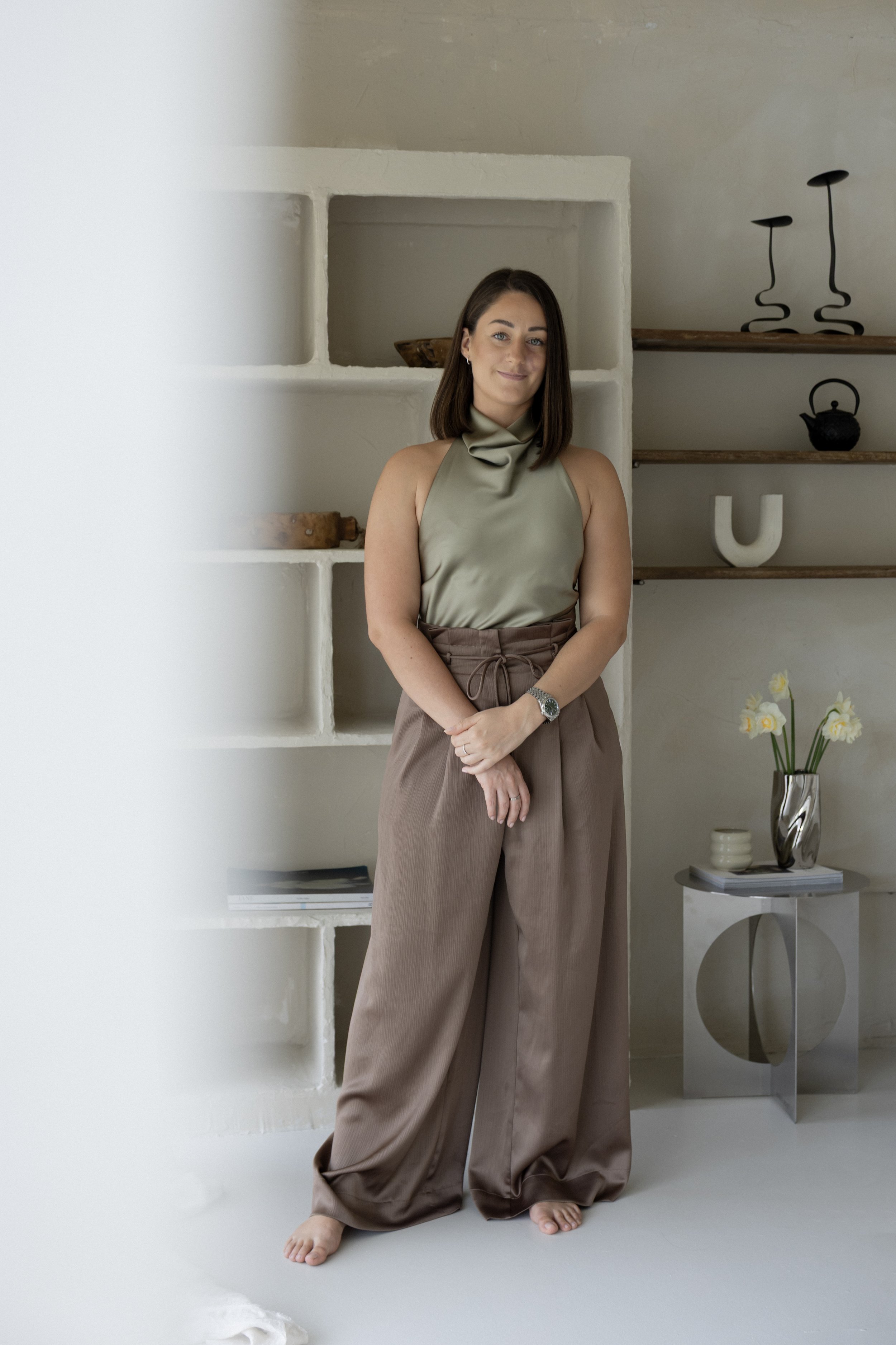 A woman with shoulder-length brown hair, wearing a beige shiny sleeveless top and loose beige pants, standing barefoot in a modern interior space with minimalist decor. She has a slight smile and is standing beside a white shelf with decorative items and a small side table with a vase of white flowers.