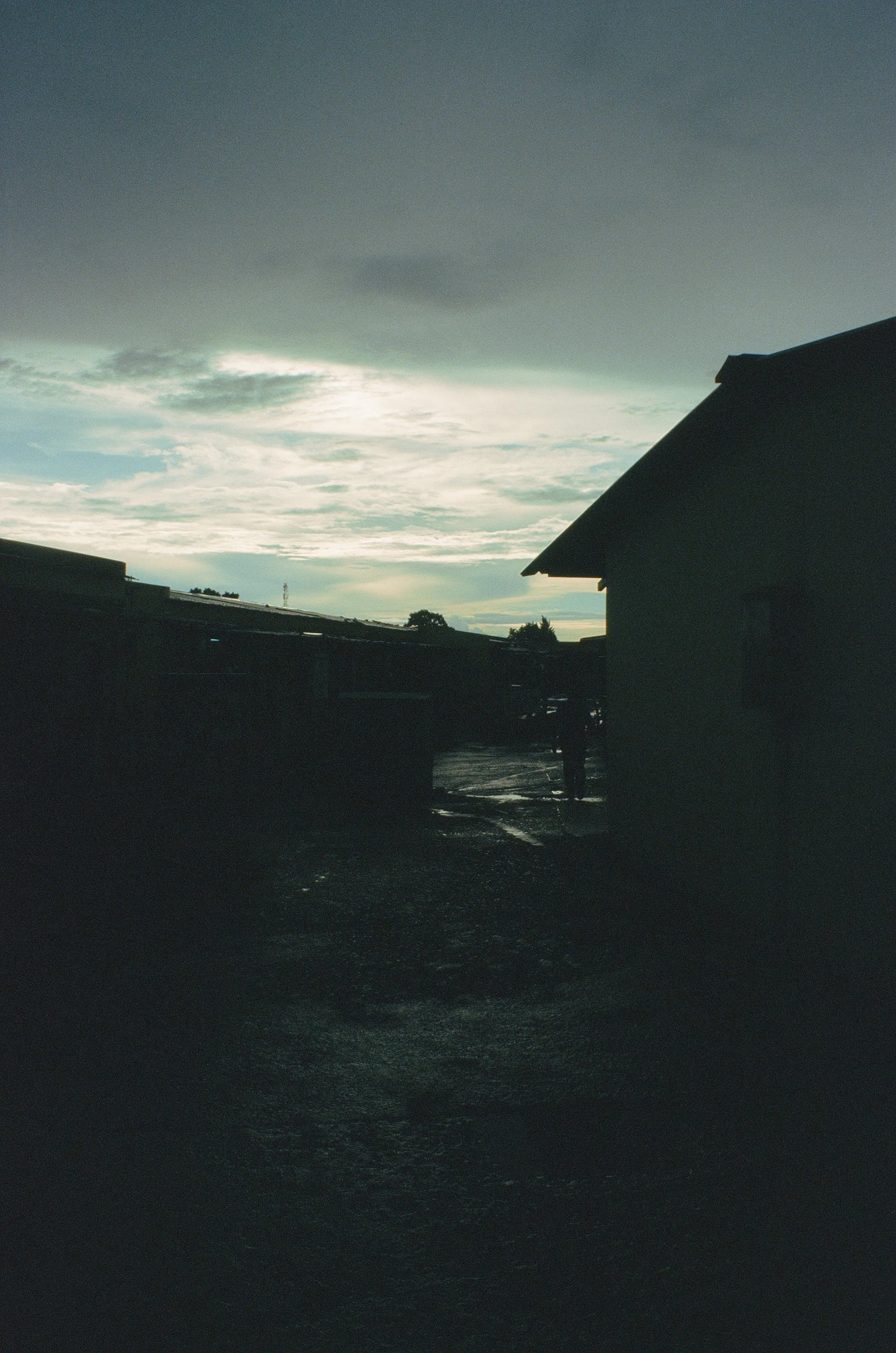 Dark silhouette of buildings and a person in the foreground with a cloudy sky and the sun peeking through at dusk or dawn.